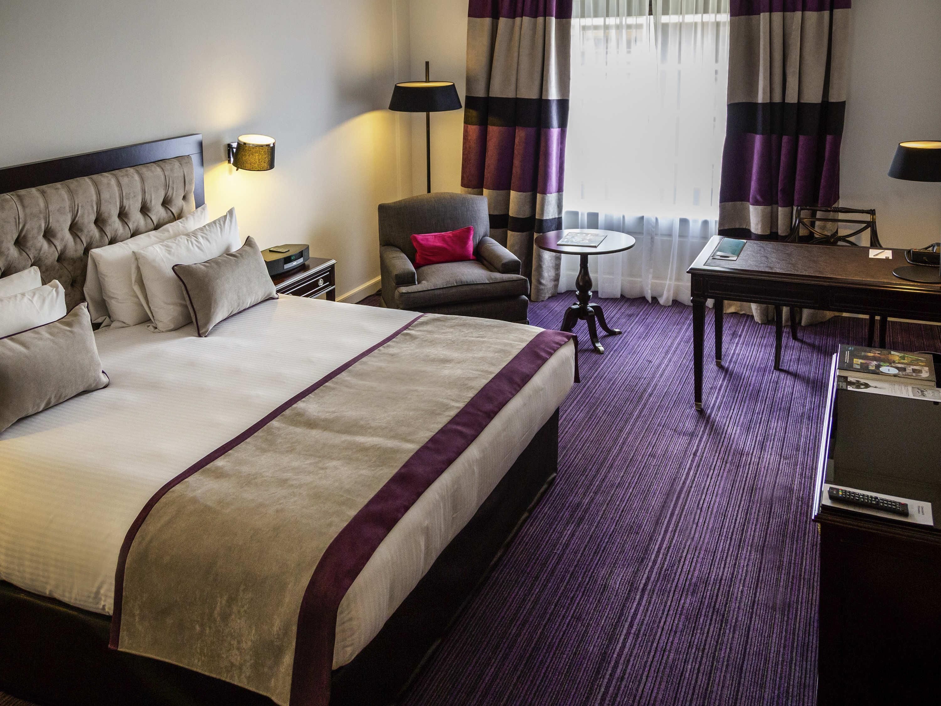 Ein Hotelzimmer mit einem großen Bett, einem Sessel, einem kleinen Tisch und einem Schreibtisch mit Lampe.
