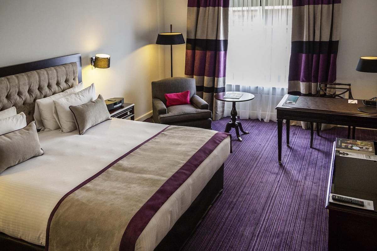 Ein Hotelzimmer mit einem großen Bett, einem Sessel, einem kleinen Tisch und einem Schreibtisch mit Lampe.