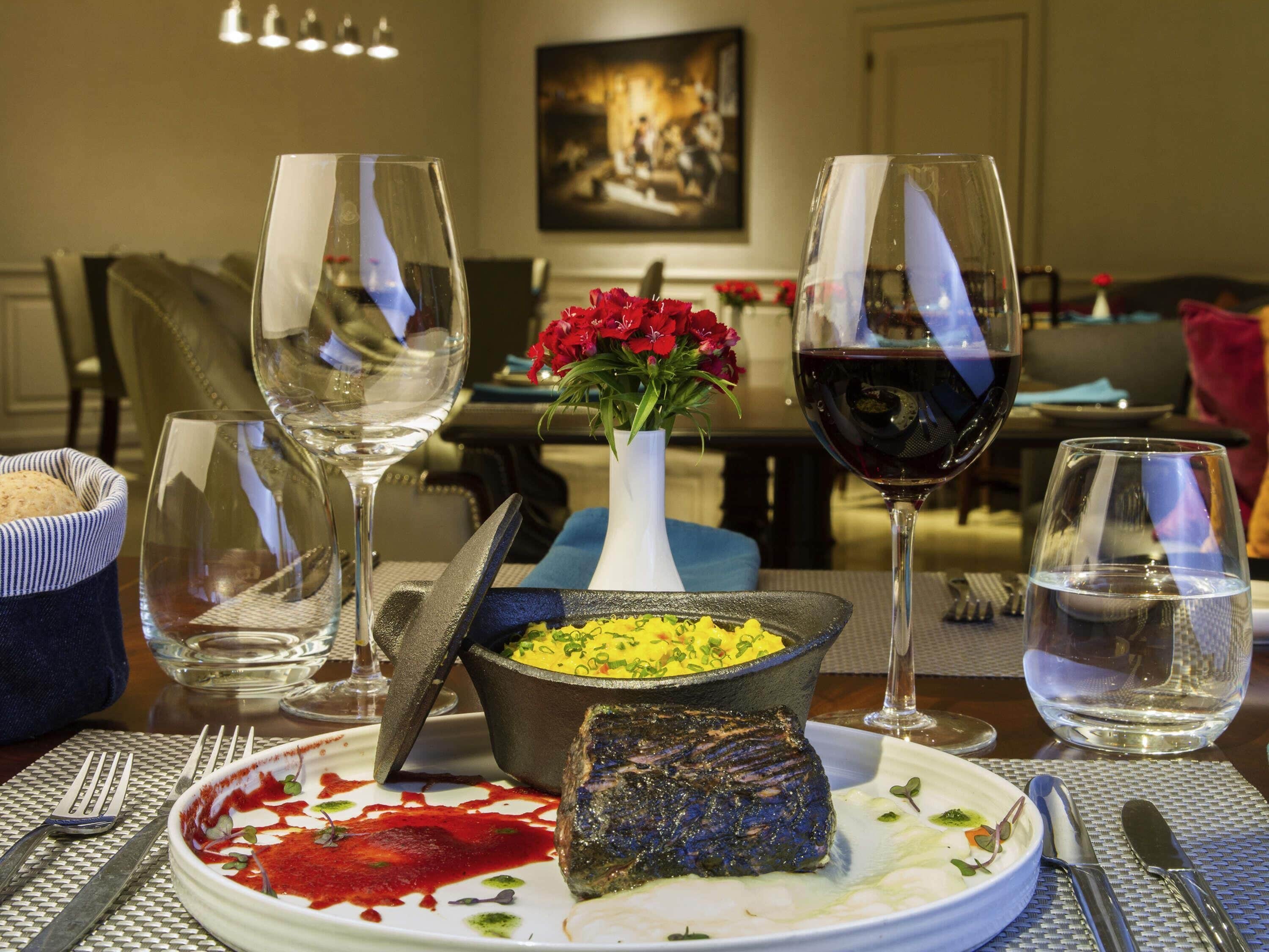 Uma mesa de jantar com um bife, uma tigela de comida amarela e um vaso de flores vermelhas. Há copos de vinho e um copo de água na mesa.