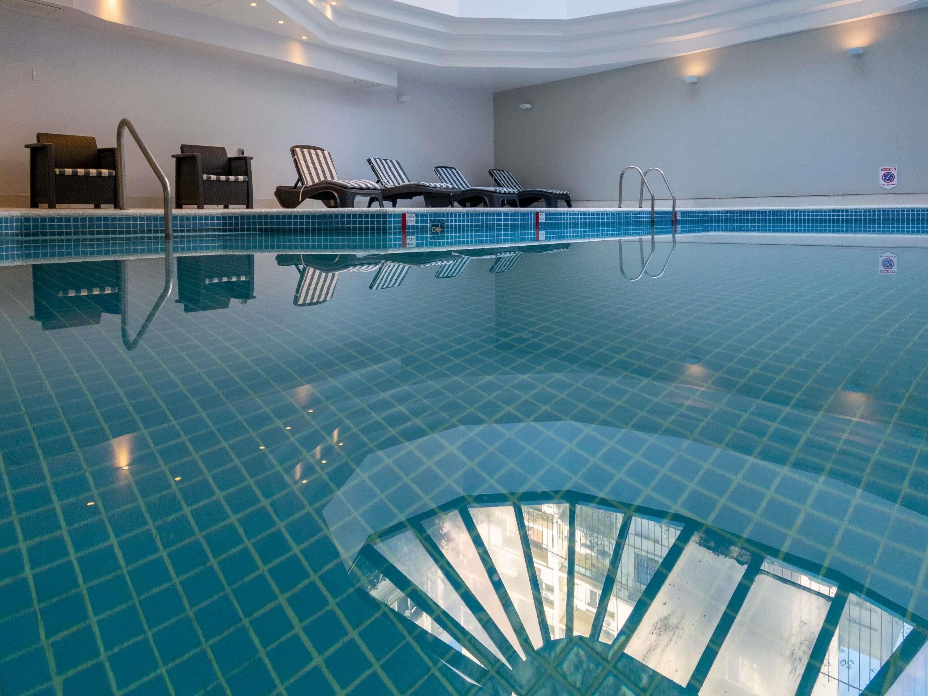 Una piscina interior cristalina con suelo de baldosas, sillones de descanso y una estructura de sombrilla.