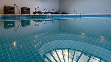 Una piscina interior cristalina con suelo de baldosas, sillones de descanso y una estructura de sombrilla.