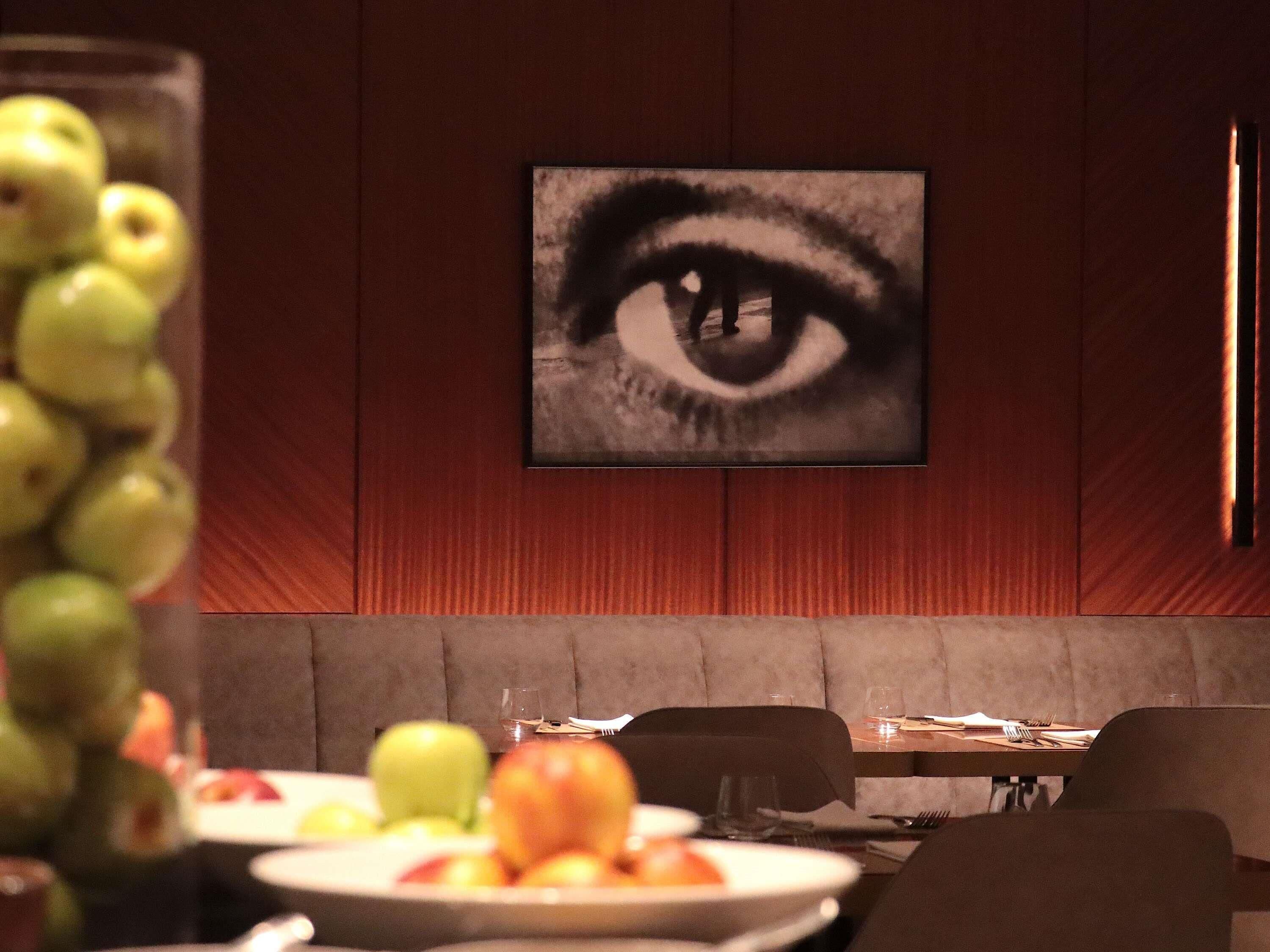 Interior de restaurante con una obra de arte de un ojo en la pared, un vaso de agua y un plato de fruta.