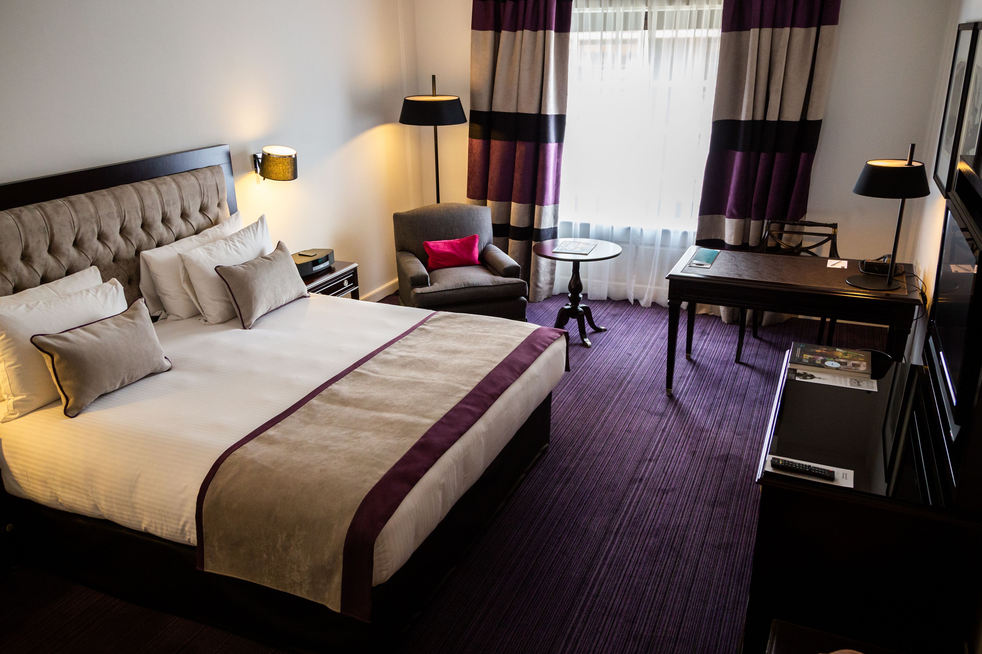 A hotel room with a large bed, a desk, a chair, a lamp, and a TV.