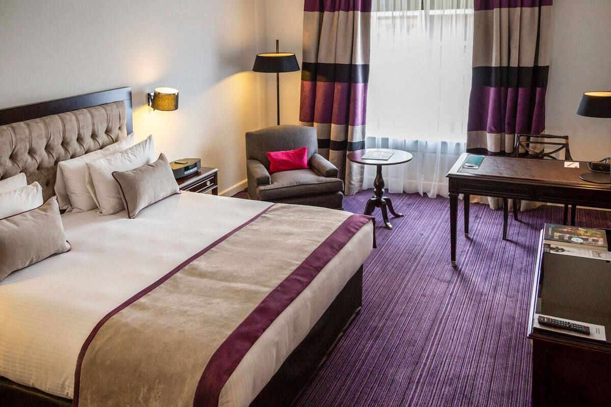 A hotel room with a large bed, a chair, a small table, and a desk.