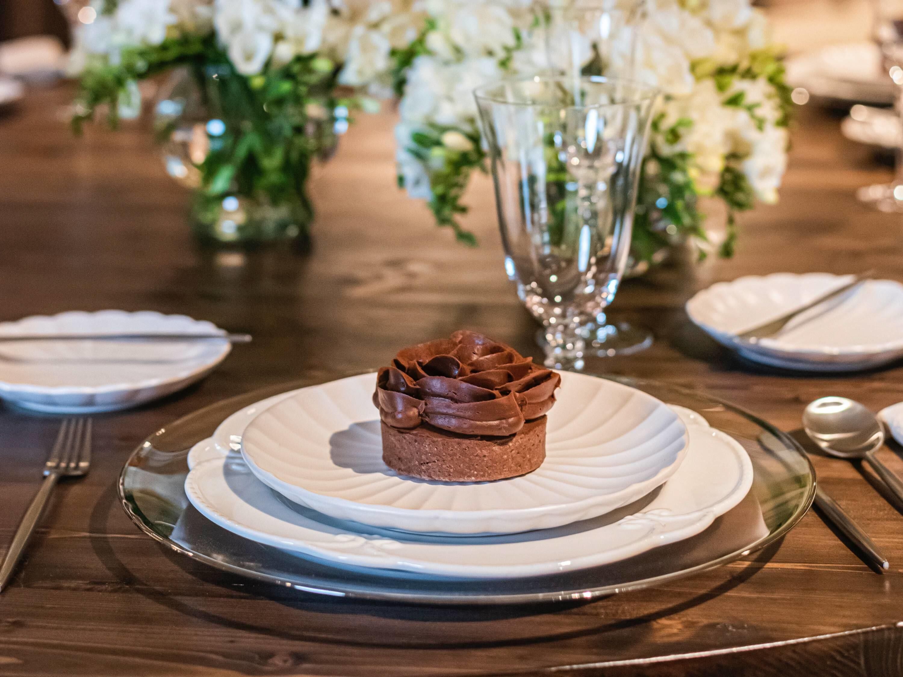 Un postre en un plato con una copa transparente debajo, rodeado de una mesa con platos, cubiertos y un jarrón con flores.