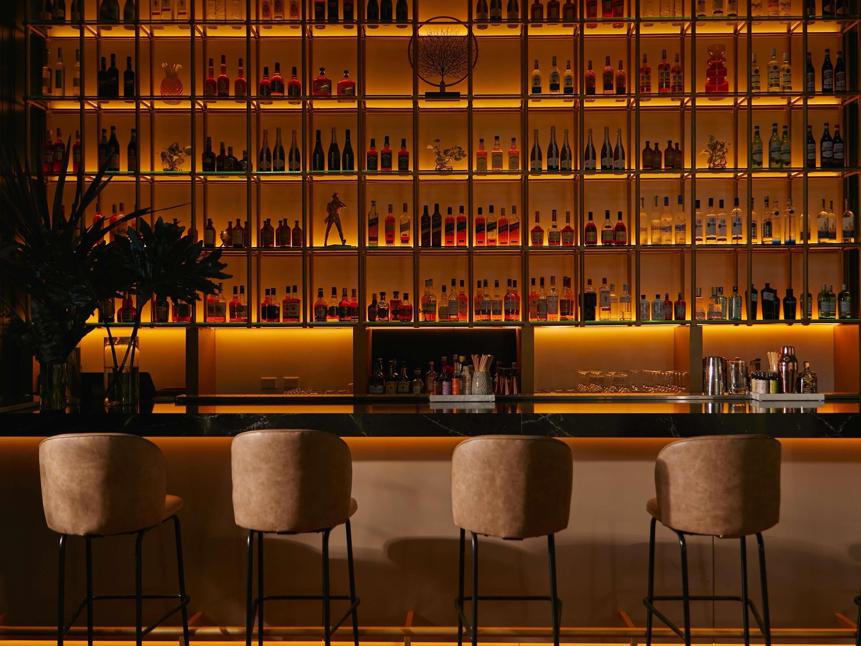 Una zona de bar con múltiples estantes llenos de botellas, una barra y tres taburetes acolchados.