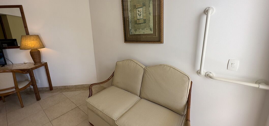 A room with a beige sofa, a wooden side table with a lamp, a framed picture on the wall, and a white handrail.