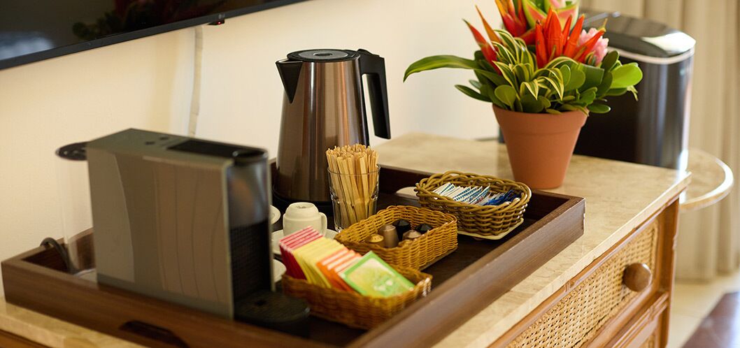 A tray with a coffee maker, a basket of snacks, a small plant, and a kettle on a wooden surface.