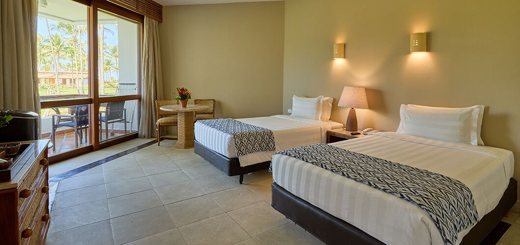 A hotel room with two beds, a small table with chairs, a TV, and a window with curtains.