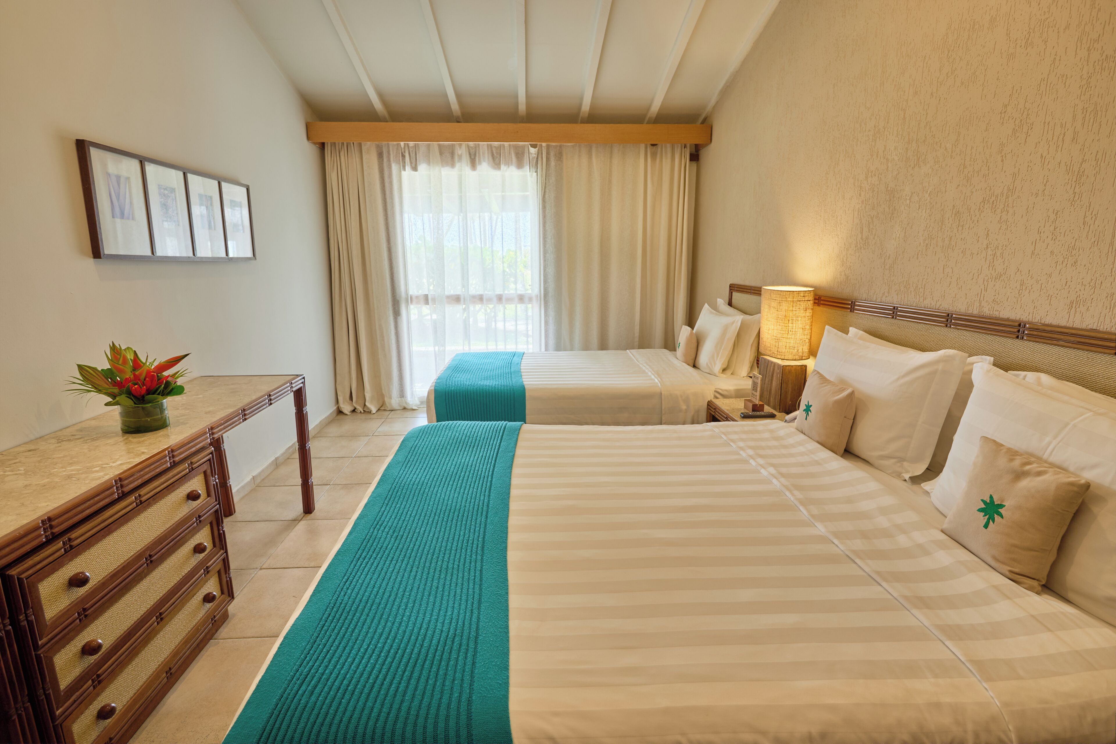 A hotel room with two beds, a wooden dresser, a desk, a lamp, and a view of the outdoors through the window.