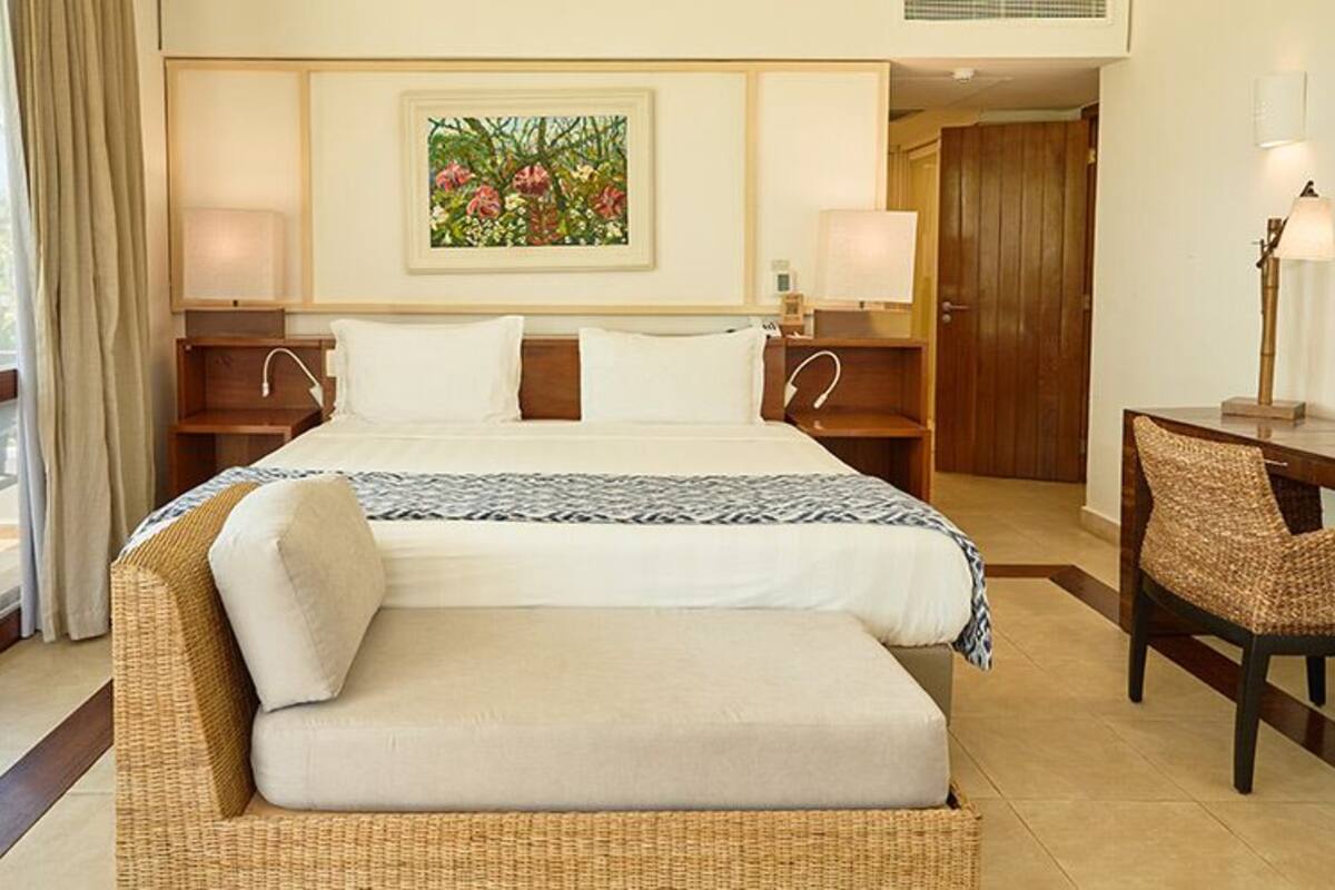 Une chambre d’hôtel avec un grand lit, un canapé en osier, un bureau avec une chaise et une vue sur les palmiers par la fenêtre.