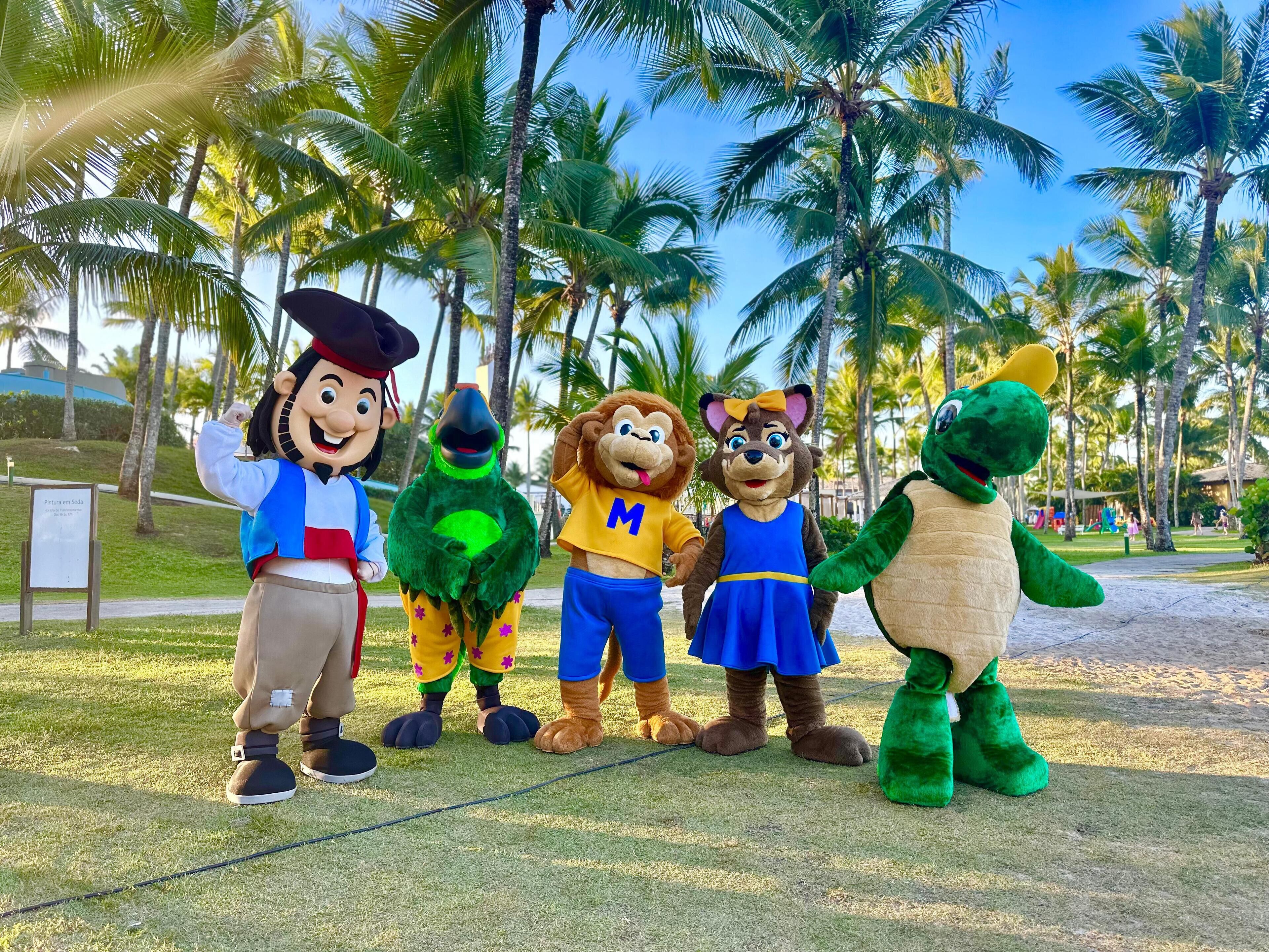 A group of mascots in costumes standing on grass with palm trees in the background.