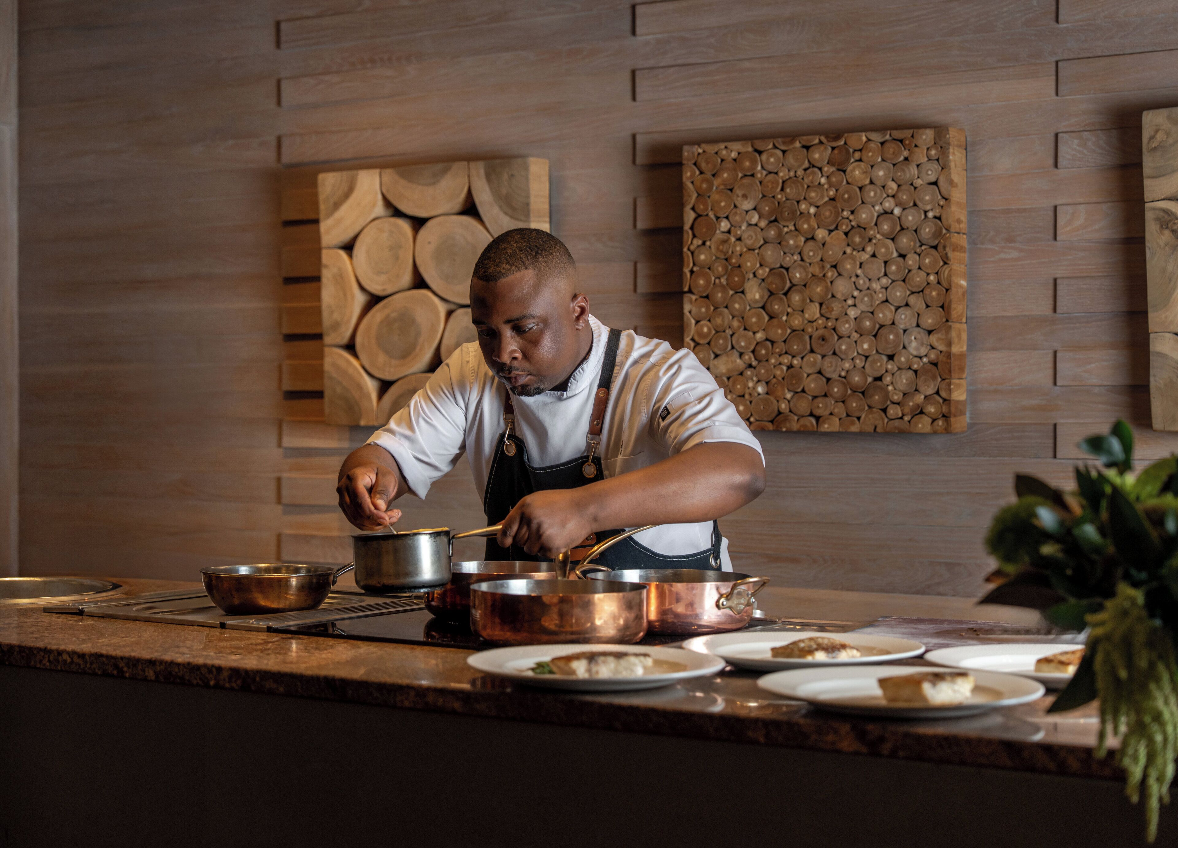 Un chef cocina en una cocina con detalles de madera en las paredes y una planta en la encimera.