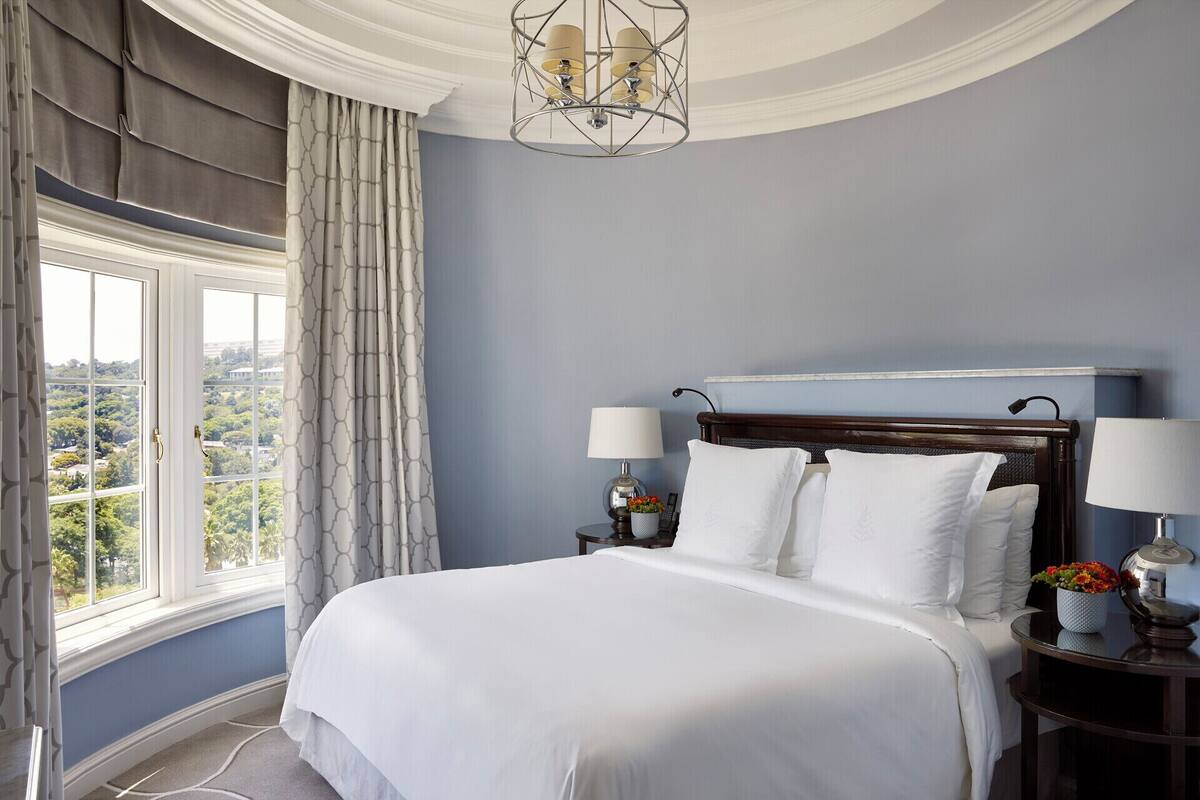 A bedroom with a large bed, two bedside tables with lamps, a view of greenery outside, and a chandelier.