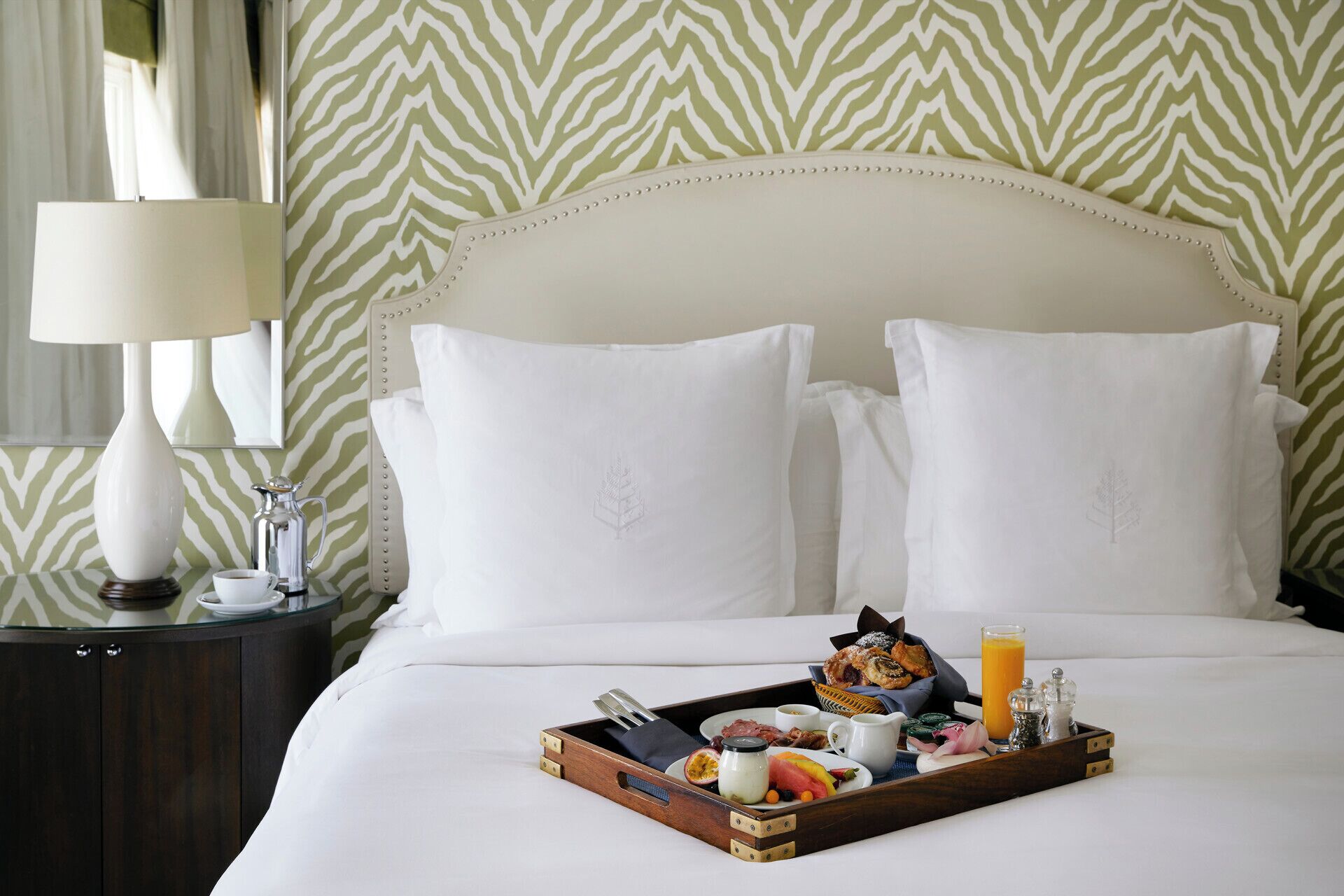 A hotel room with a bed, pillows, a tray with breakfast, and a nightstand with a lamp.