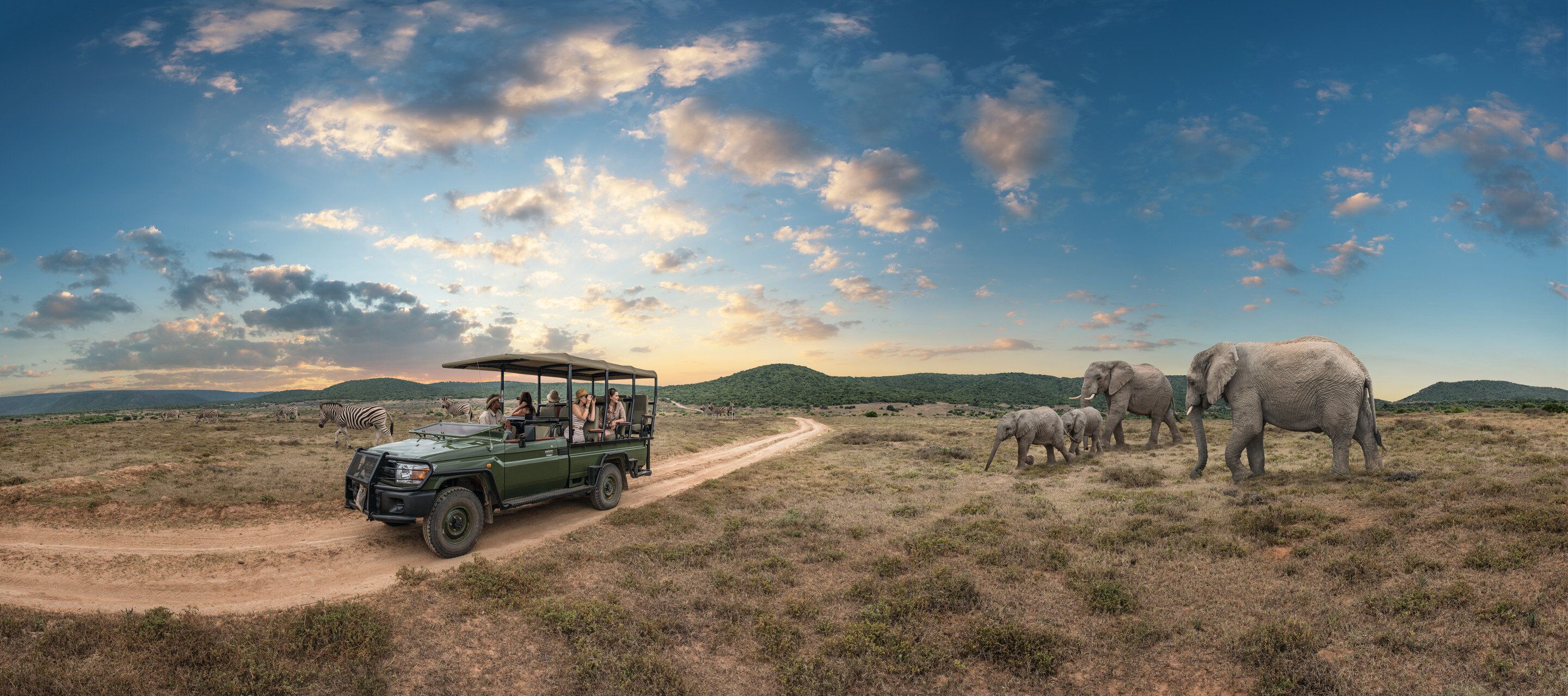 Un vehículo safari con pasajeros observando fauna silvestre, incluyendo elefantes y cebras, en un paisaje de sabana.