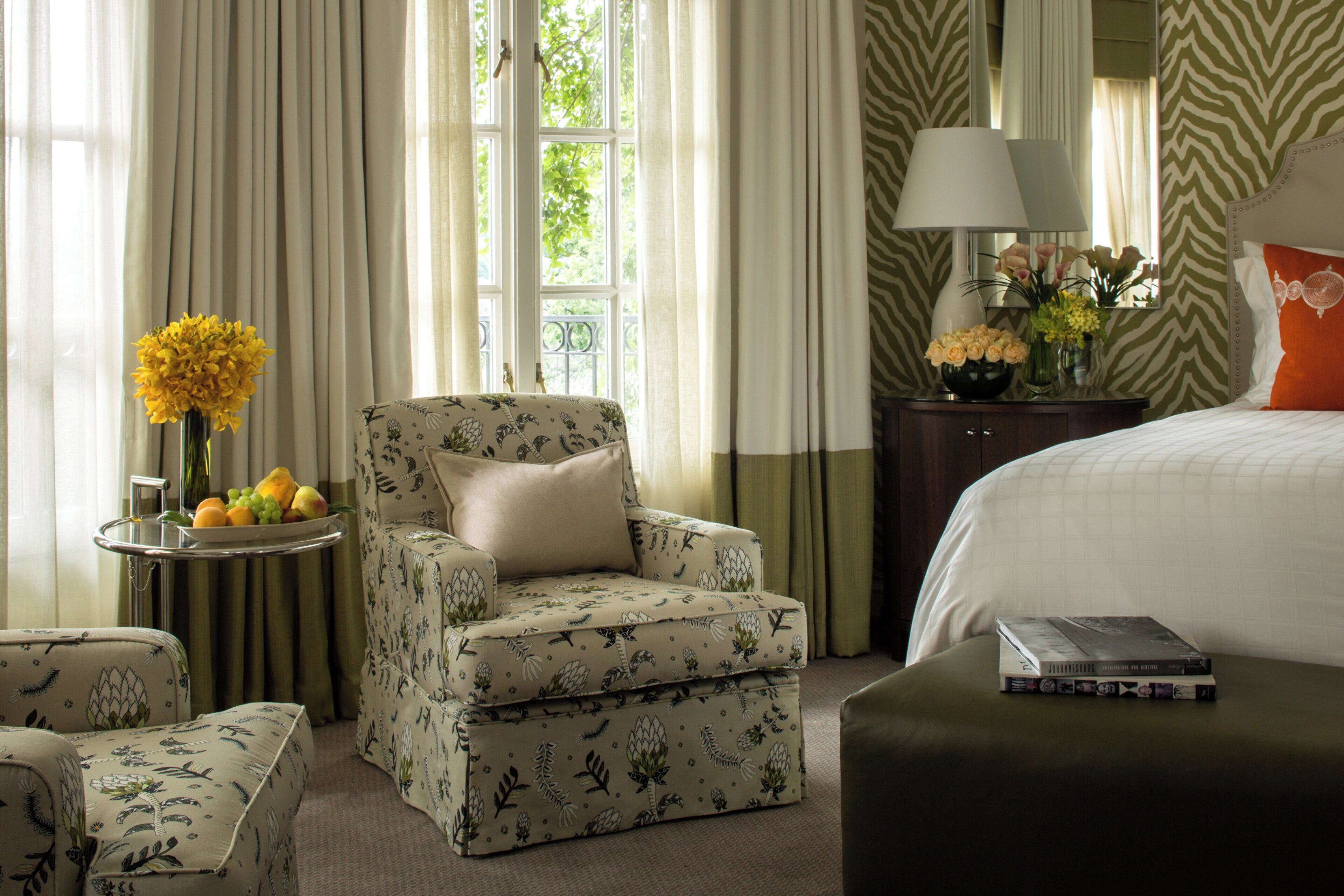 A hotel room with a bed, a chair, a small table with fruit and flowers, and a lamp.