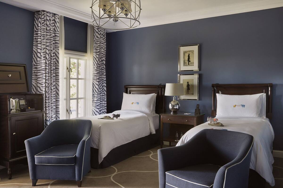 A hotel room with two beds, a desk, a chair, a lamp, and a framed picture on the wall.