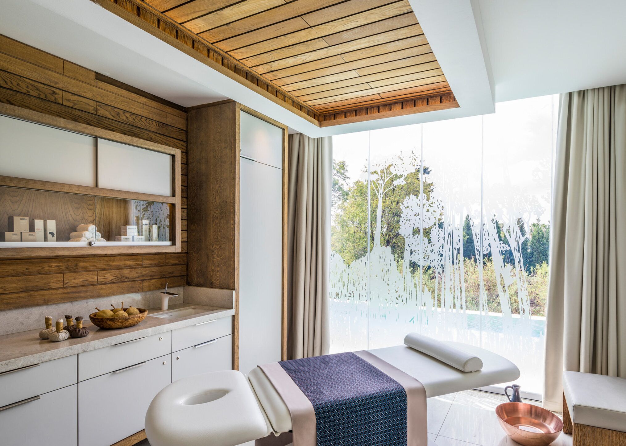 Un spa moderno con vigas de madera en el techo, un amplio ventanal con vistas a árboles, una camilla de masaje y un mostrador con artículos de aseo.