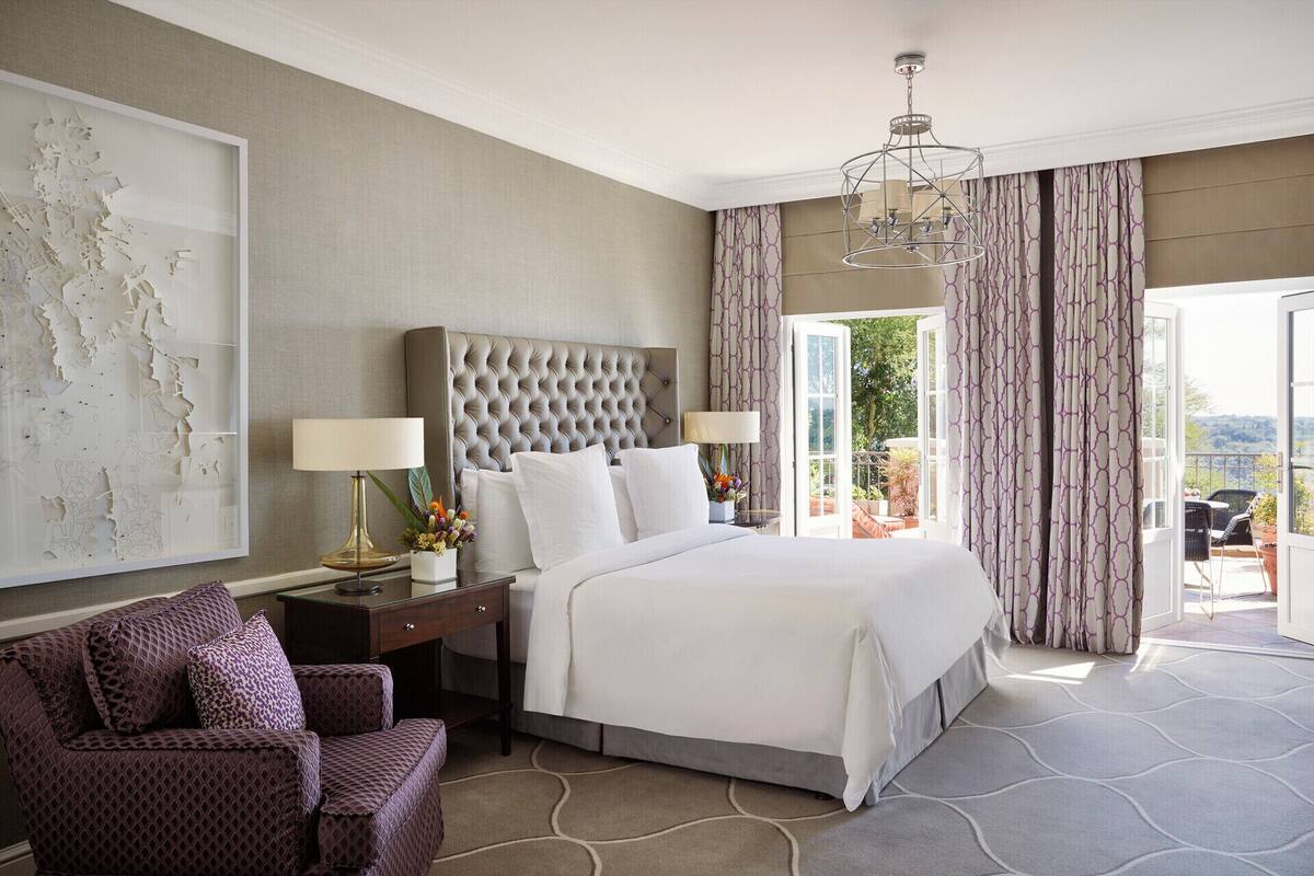 A hotel room with a large bed, two bedside tables, a chair, a large window with curtains, and a view of the outdoors.