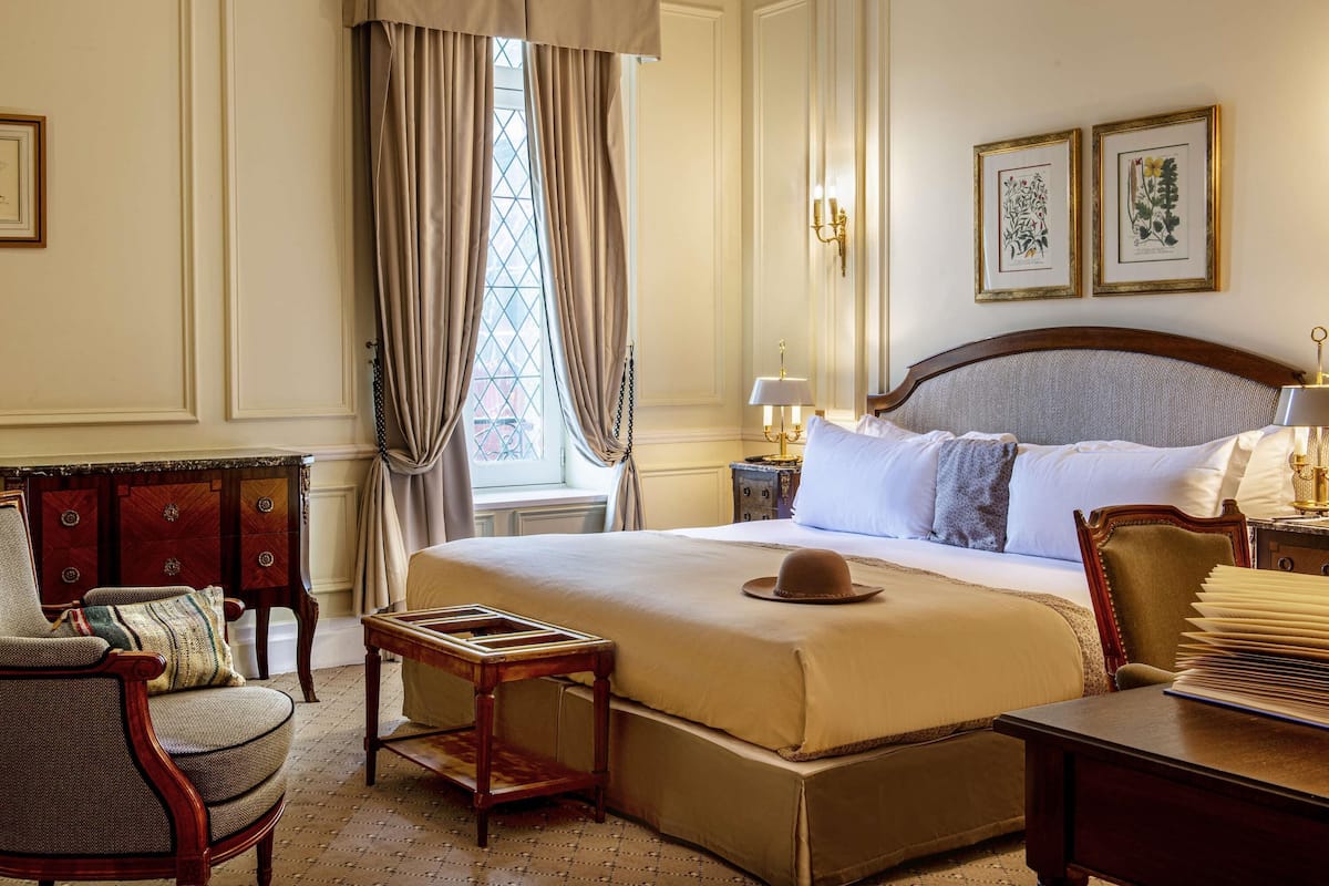 Ein klassisches Hotelzimmer mit einem großen Bett, einem Holznachttisch, einem Sessel und einem Schreibtisch mit Lampe.