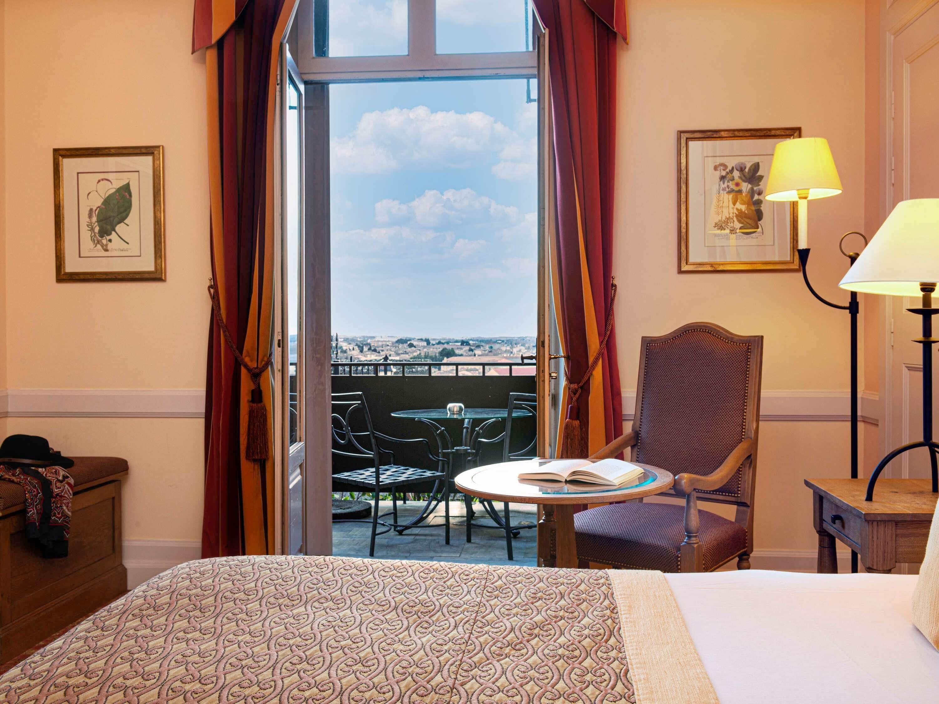 A hotel room with a bed, a chair, a table with a book, a lamp, and a balcony with a view.