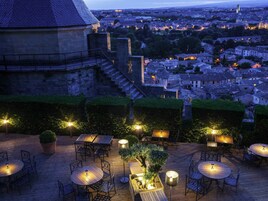 Un patio al aire libre con mesas y sillas, un edificio de piedra al fondo y vista a la ciudad.