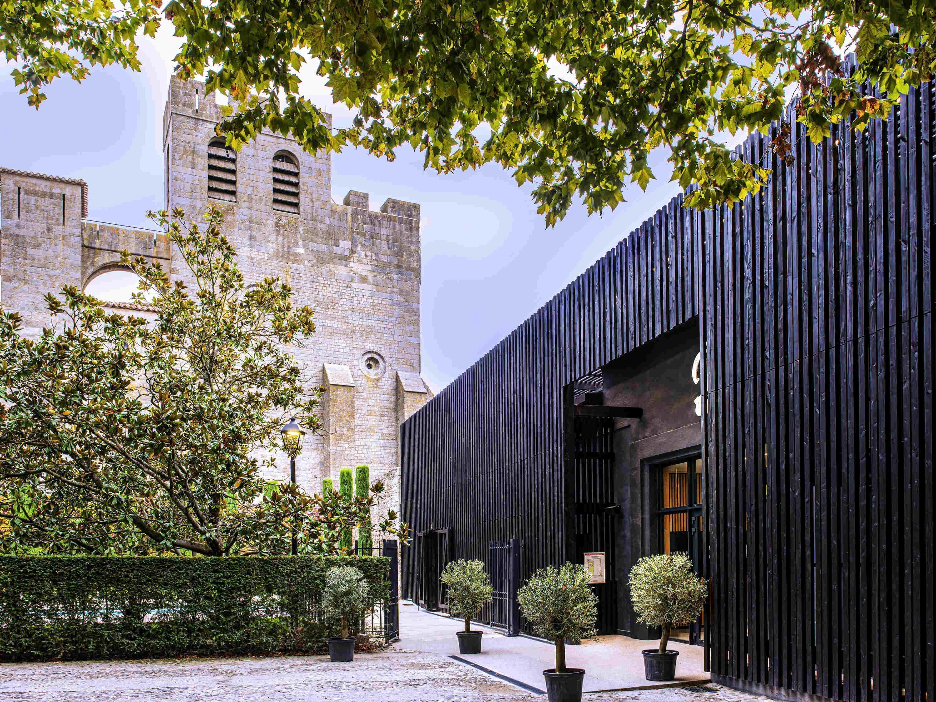 Un patio con un edificio histórico de piedra, una estructura moderna de color negro y abundante vegetación.