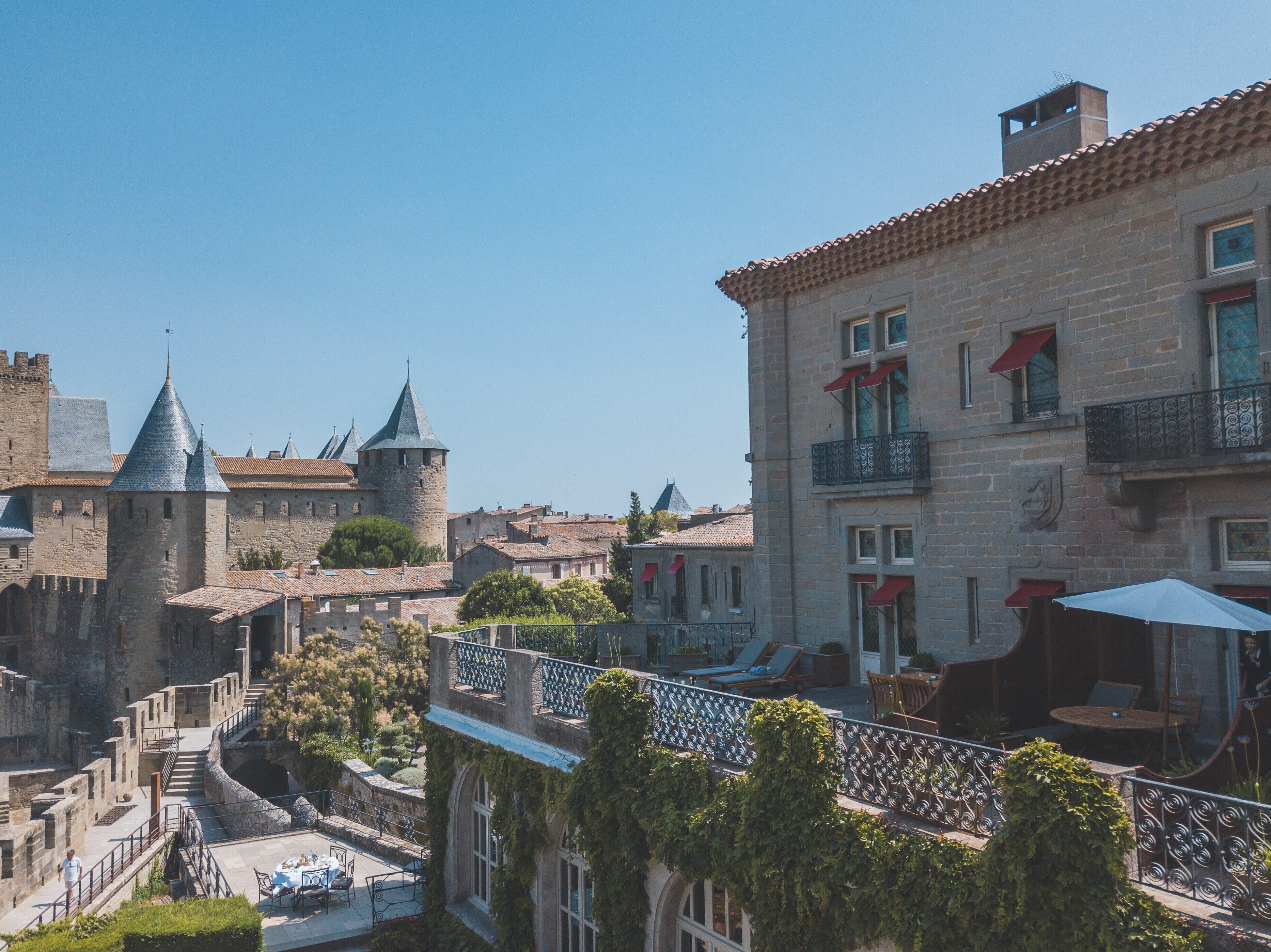A courtyard with a stone tower, a building with a balcony, and a view of a castle in the background.