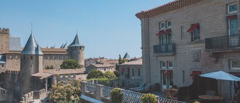 A courtyard with a stone tower, a building with a balcony, and a view of a castle in the background.