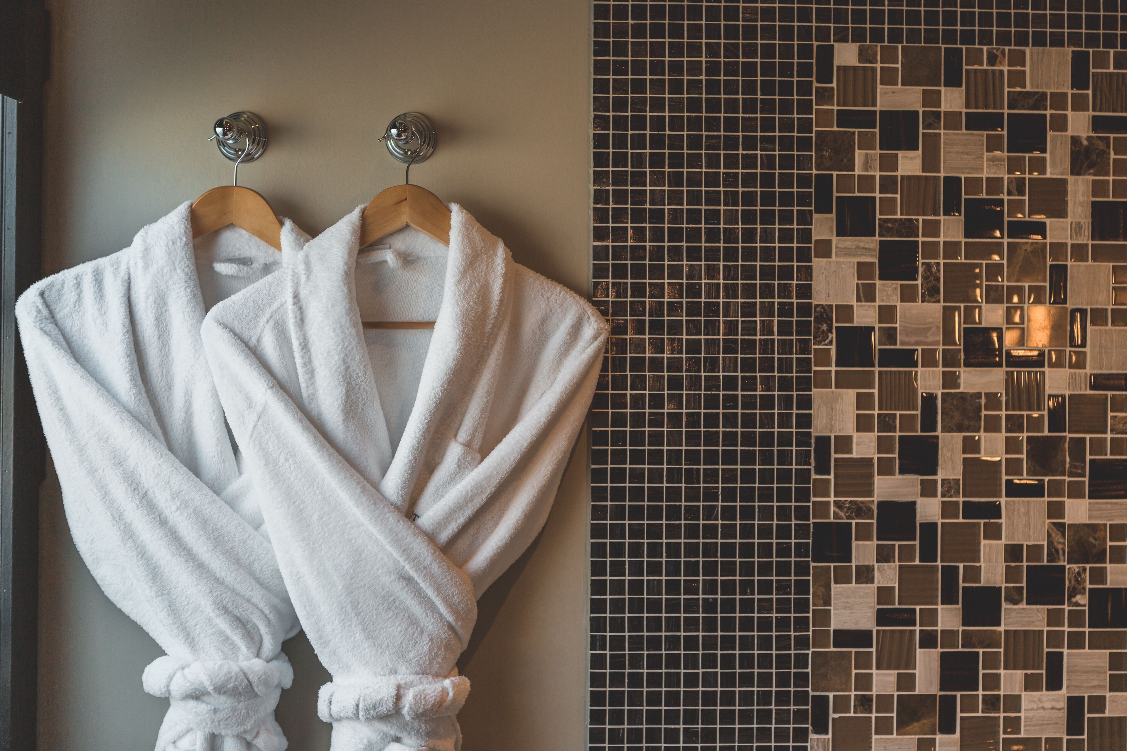 Two white bathrobes hanging on a wooden hanger against a wall with a mosaic tile pattern.