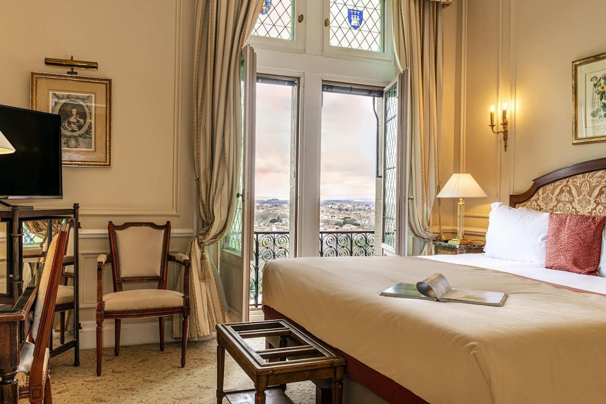 Habitación de hotel con cama, escritorio, silla, televisor y vistas de la ciudad a través de amplias ventanas.