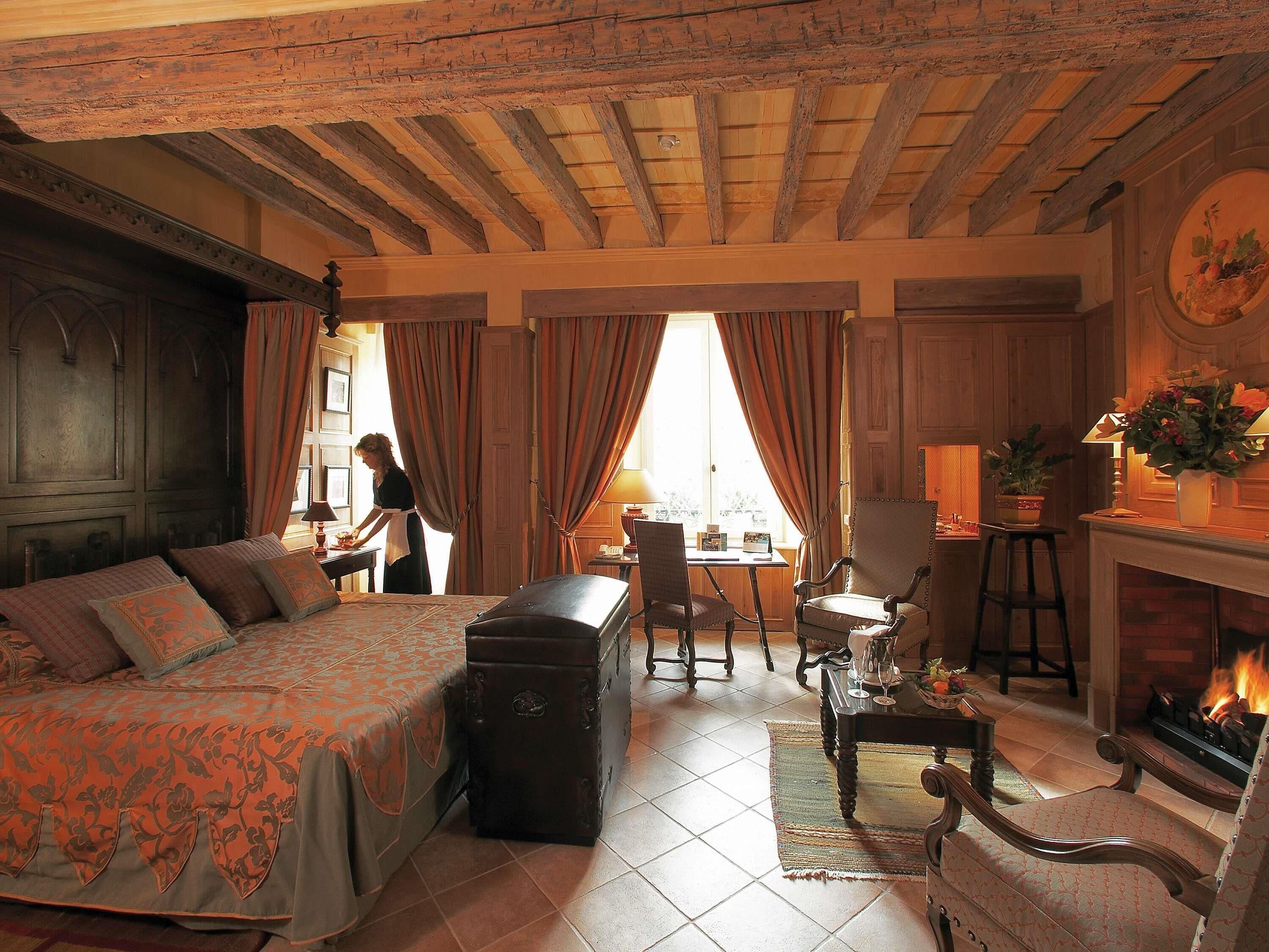 A traditional room with a bed, a desk, a chair, a fireplace, and a window with curtains.
