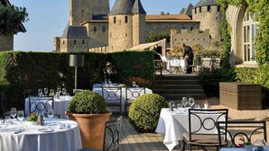 Un espacio de comedor al aire libre con mesas preparadas para los huéspedes, un castillo al fondo y un seto en primer plano.