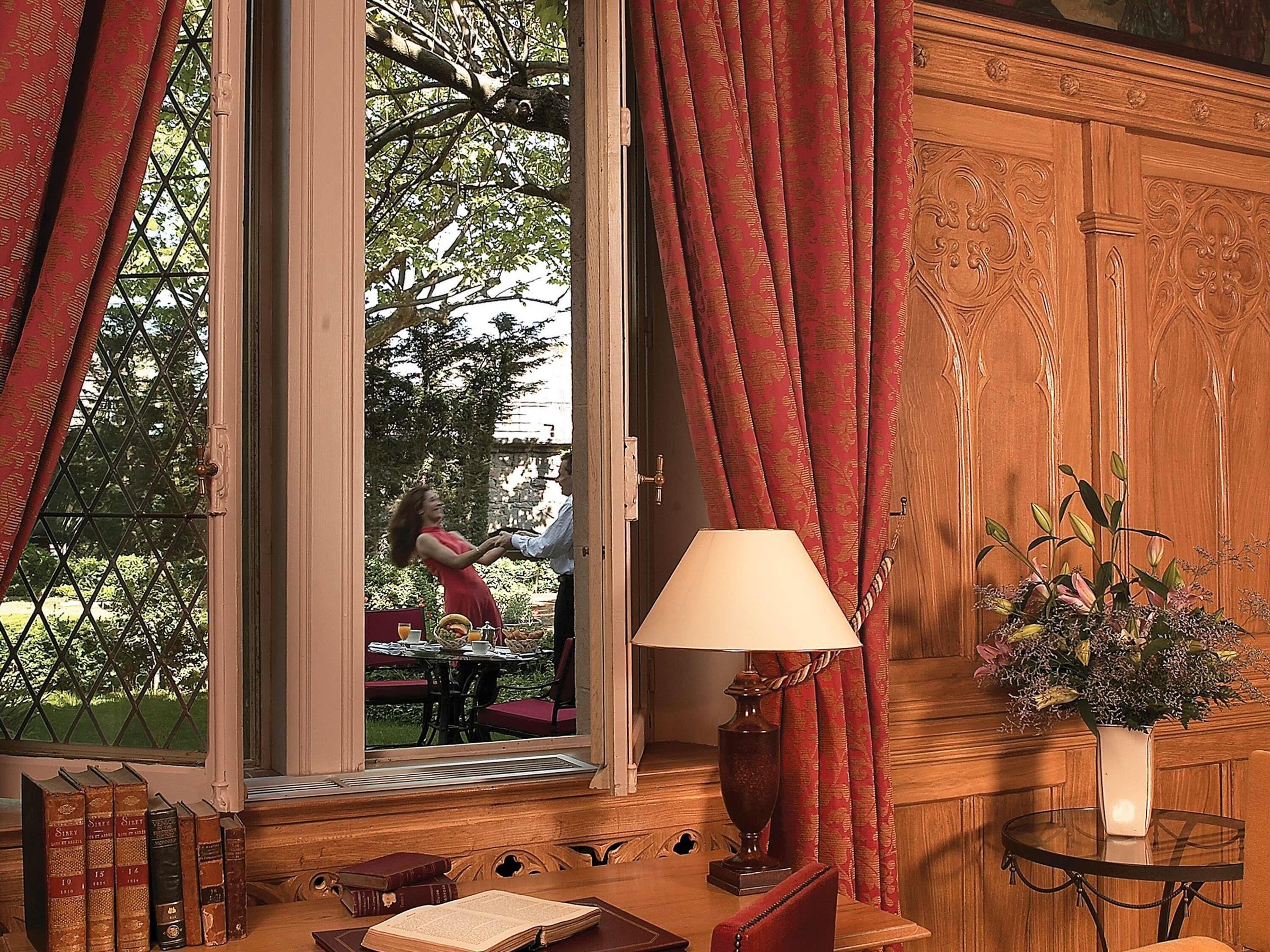 A room with a large window, a table with a lamp, a vase with flowers, and books on a shelf.