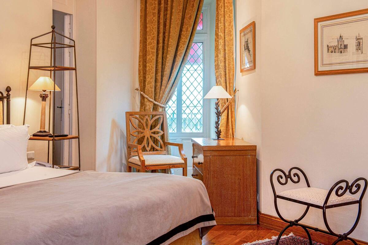 Un dormitorio con cama, una silla, un escritorio, una lámpara, una ventana con cortinas y un cuadro en la pared.