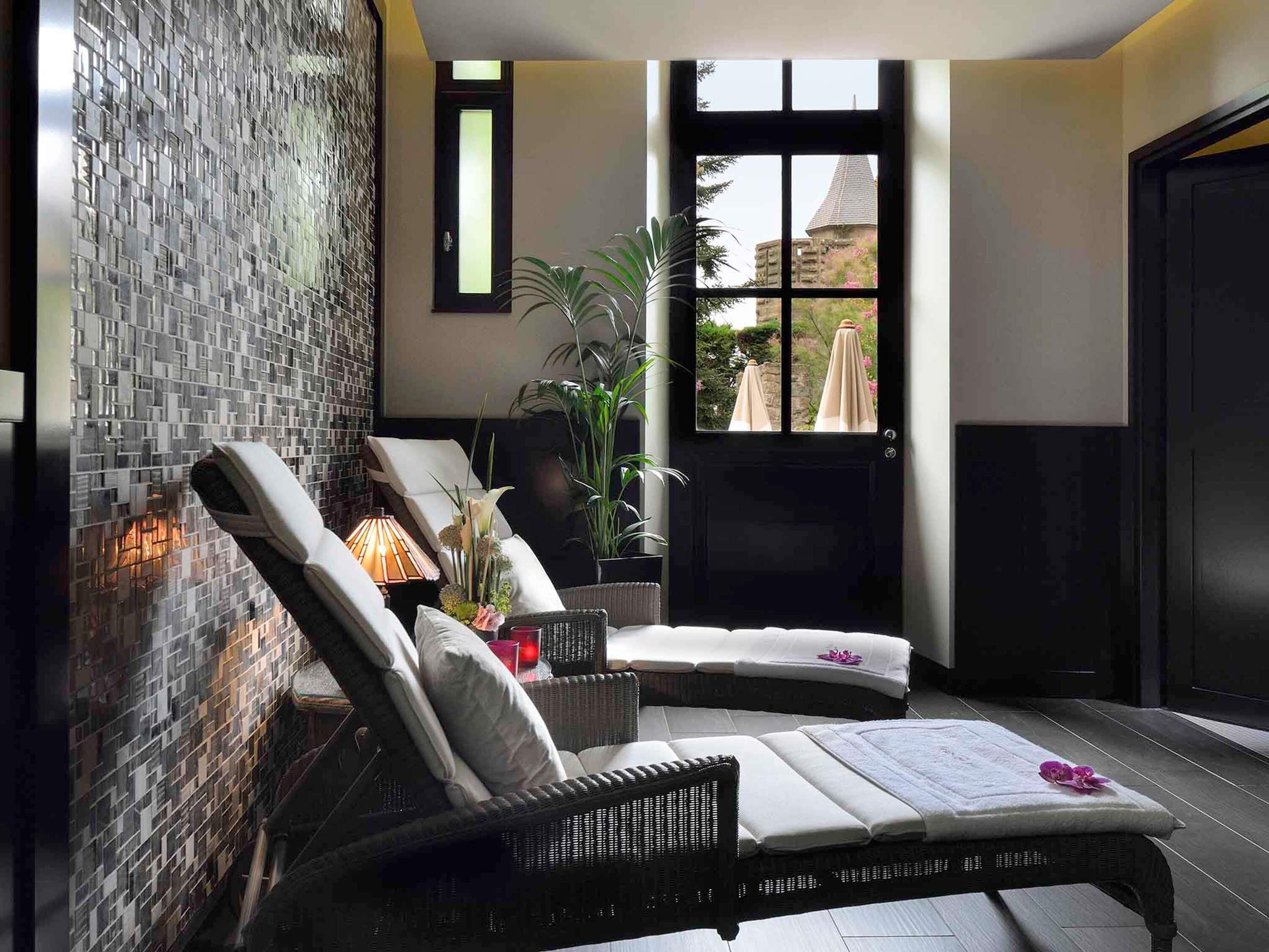 Un espace spa comprenant un fauteuil, une table de massage, une plante en pot et une vue sur un bâtiment par le biais d’une fenêtre.