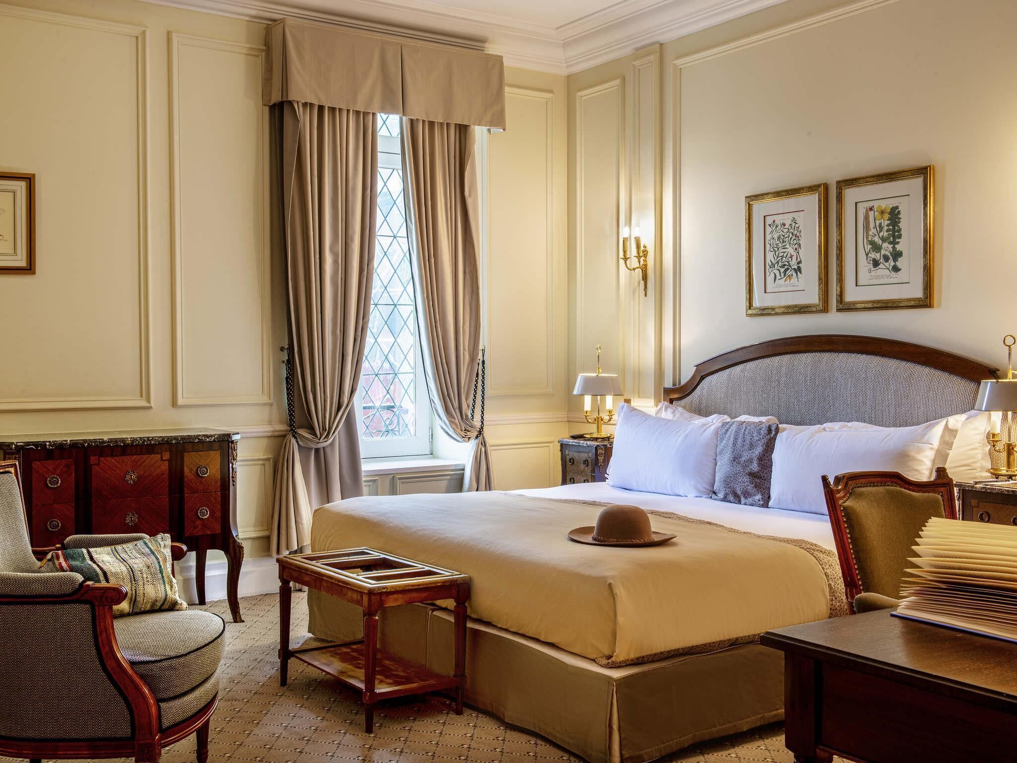 Una habitación de hotel clásica con una cama grande, una mesita de noche de madera, una silla y una ventana con cortinas.