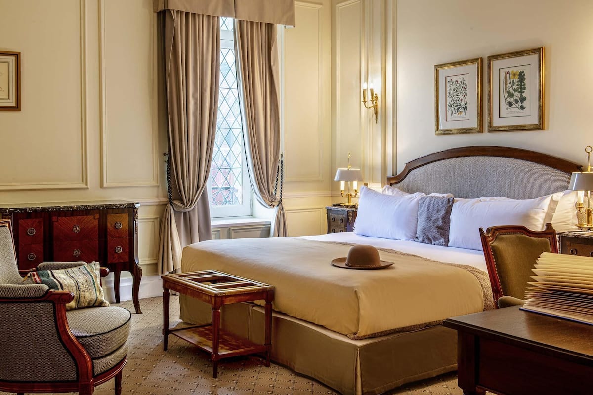 A classic hotel room with a large bed, a wooden nightstand, a chair, and a window with curtains.