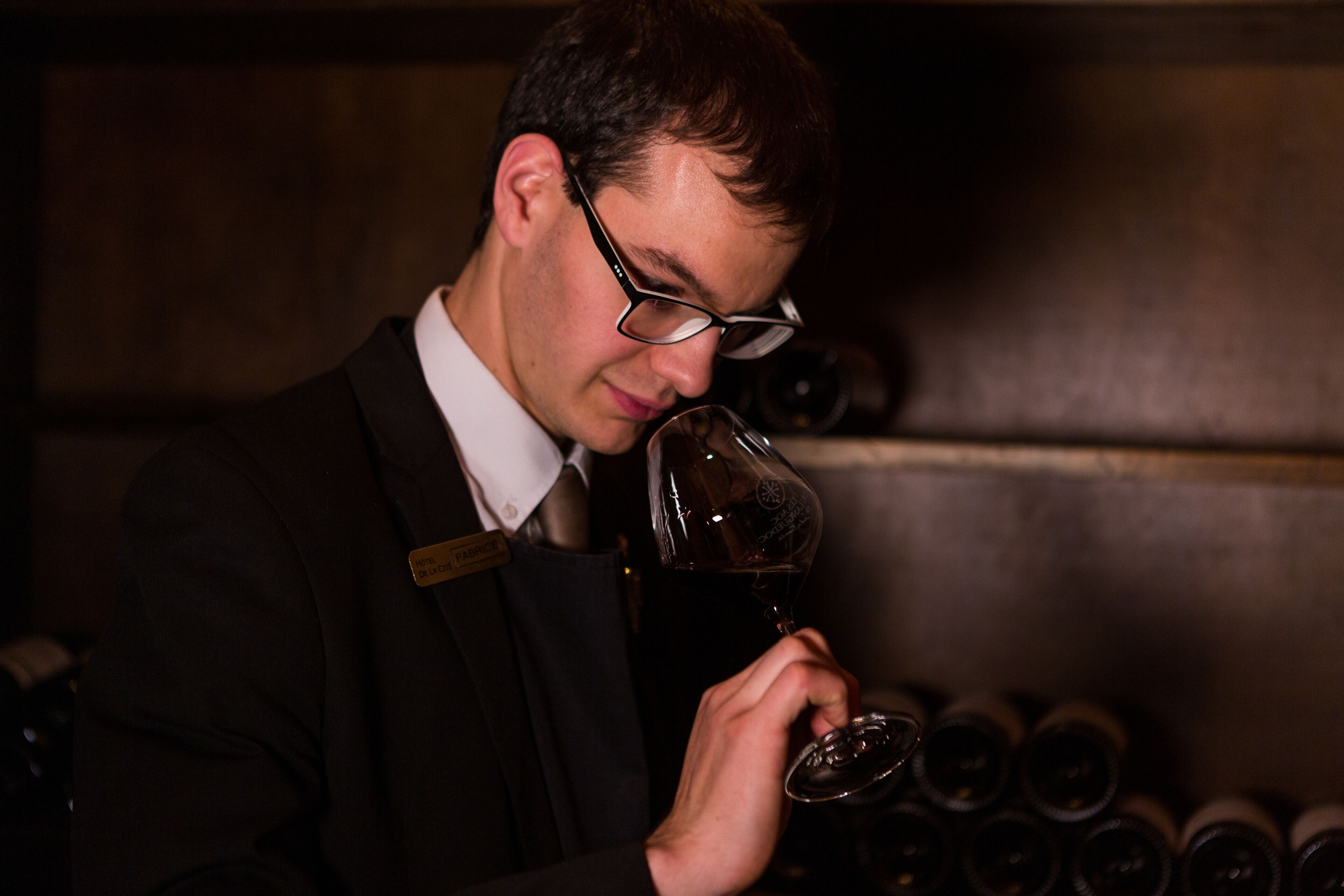 A man in a suit holding a wine glass, standing in front of a wall with wine bottles.