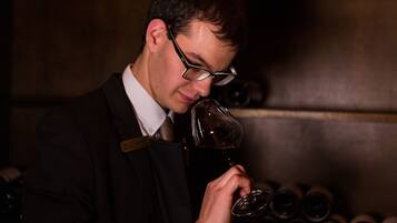 A man in a suit holding a wine glass, standing in front of a wall with wine bottles.