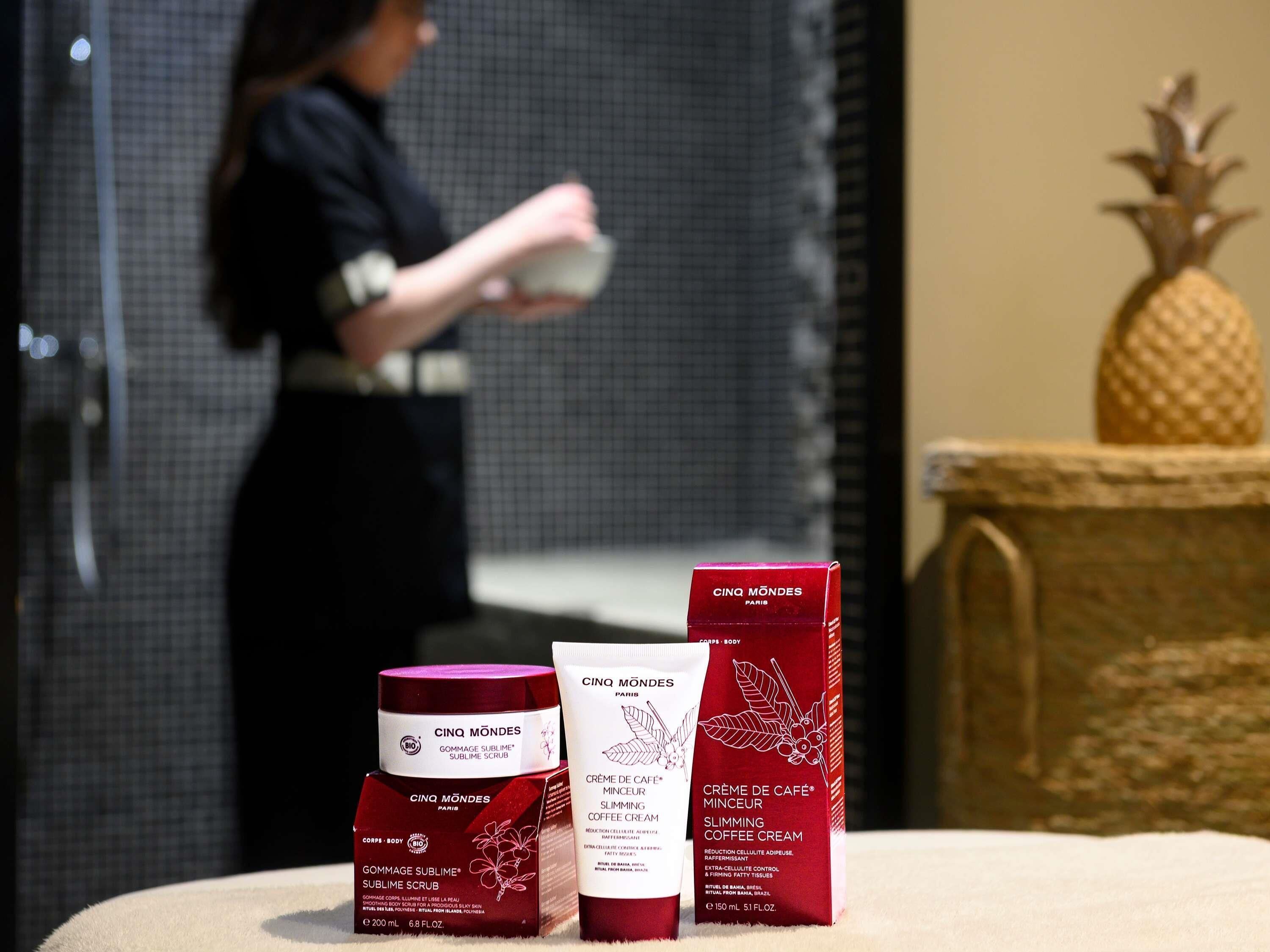 A bathroom with a shower, a woman in the background, and three boxes of Cinq Mondes Paris products on a counter.