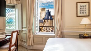 Habitación de hotel con un ventanal grande, una cama, un escritorio con libros y un cuadro en la pared.