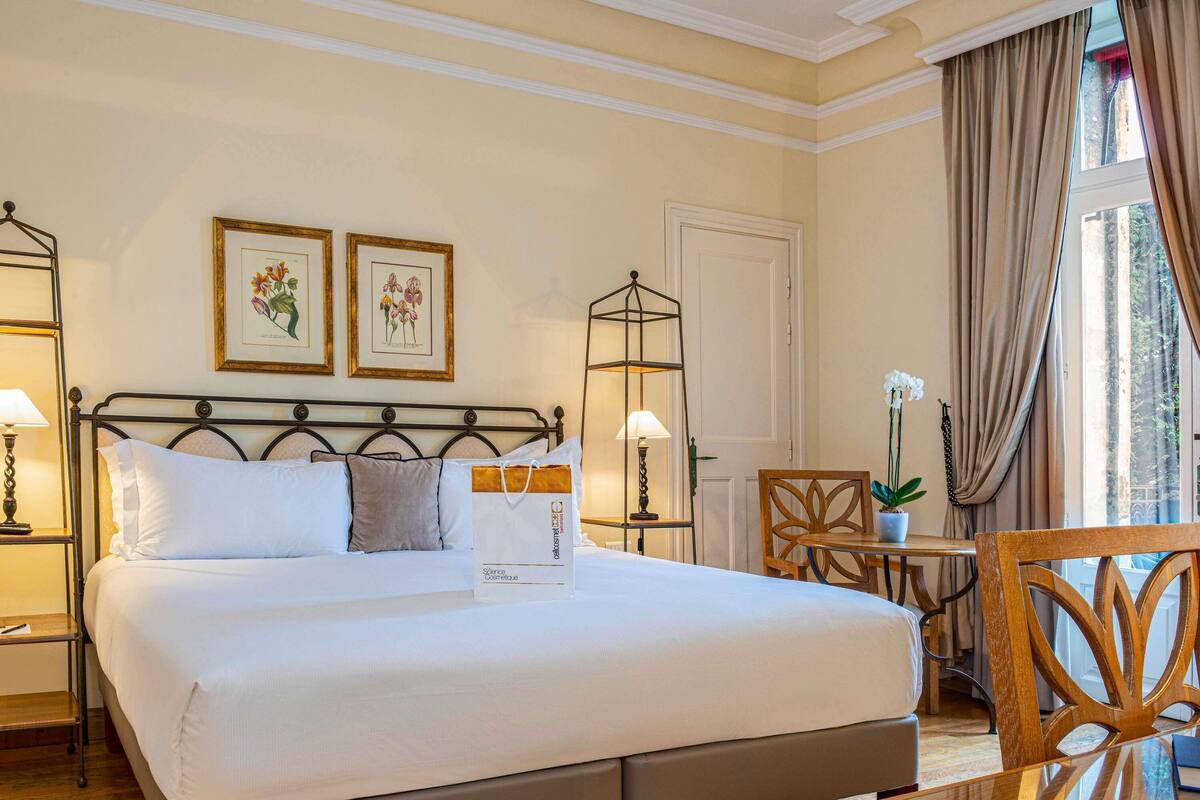 Una cama bien tendida con almohadas, una mesita de noche con lámpara, una silla y una pequeña mesa con una planta en maceta.