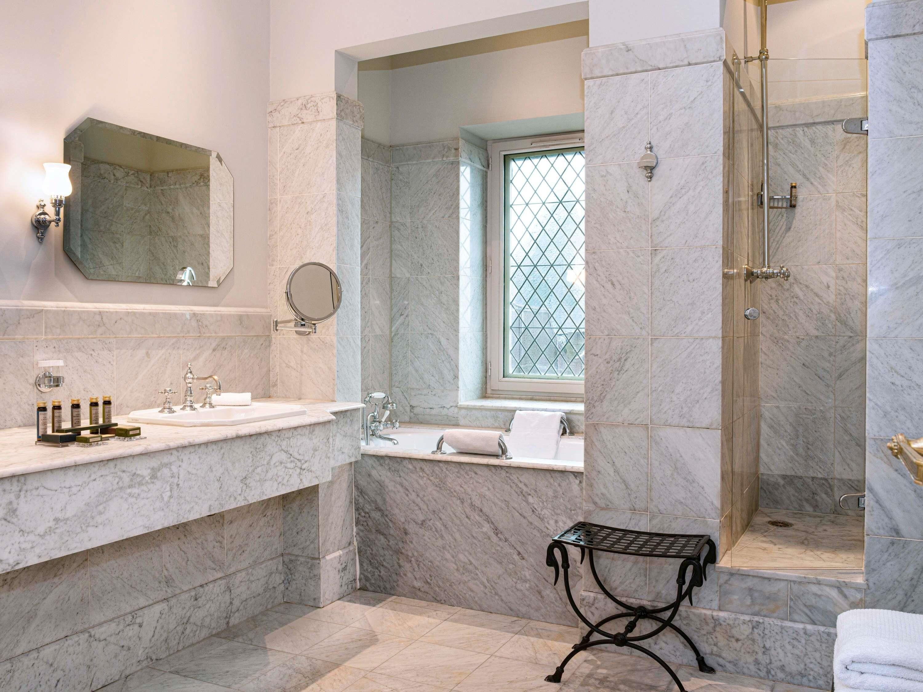 Un baño con paredes de mármol, un espejo grande, una ventana, ducha y lavabo con artículos de aseo.
