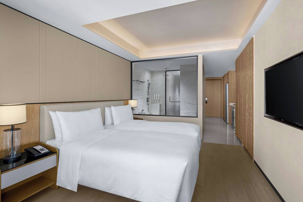 Ein modernes Hotelzimmer mit einem gro ßen Bett, einem Nachttisch mit Lampe, einem Fernseher und einem sichtbaren Duschbereich hinter einer Glastür.