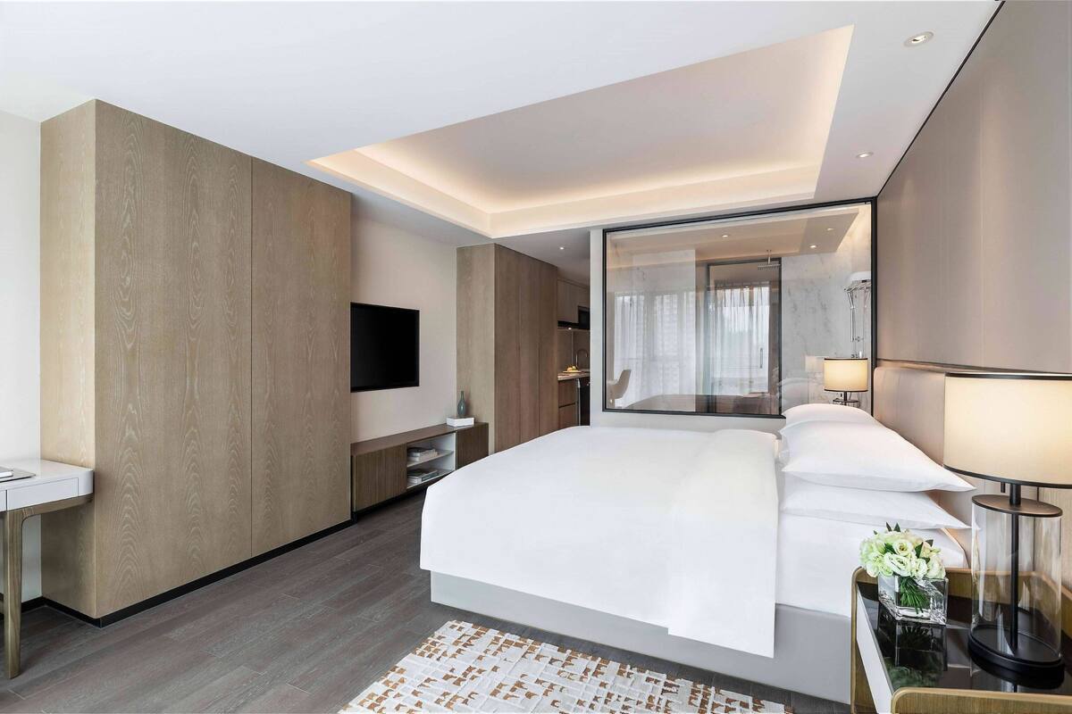 Ein modernes Hotelzimmer mit einem großen Bett, einem Flachbildfernseher, einem Nachttisch mit Lampe und einem durch eine Glastür einsehbaren Badezimmer.