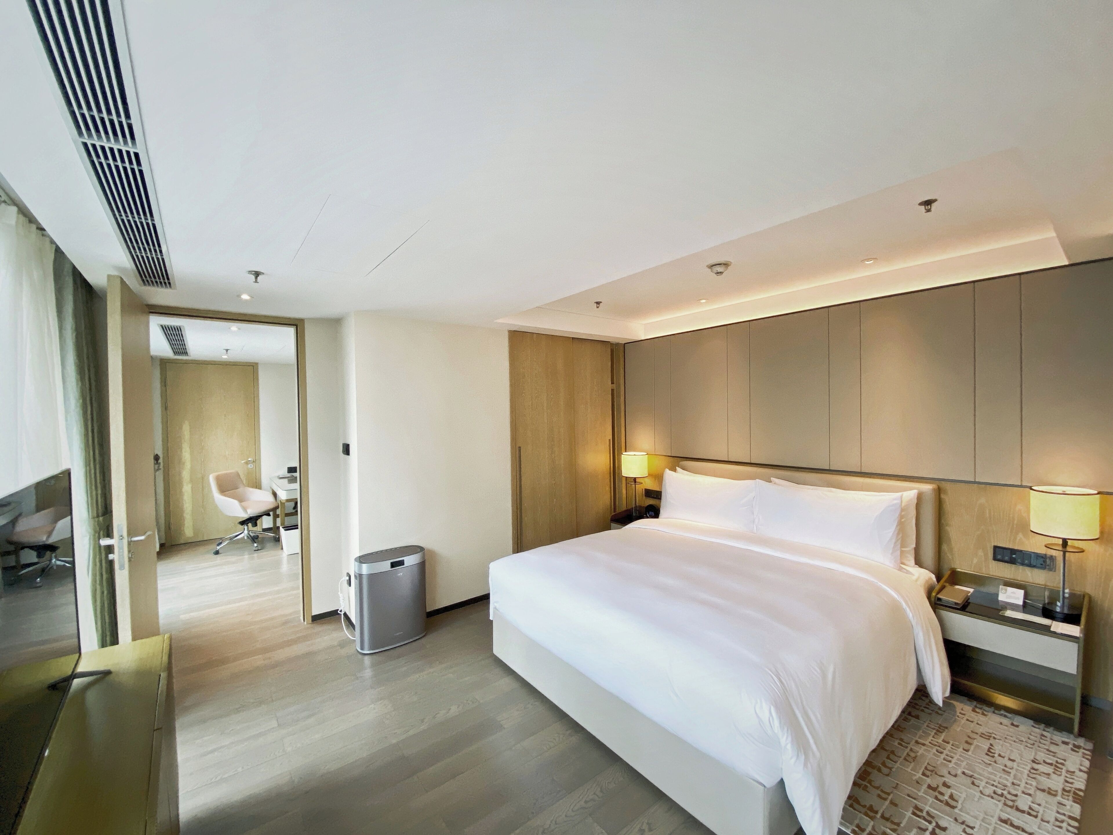 A modern hotel room with a large bed, bedside tables with lamps, a waste bin, and a view of another room with a chair and desk.