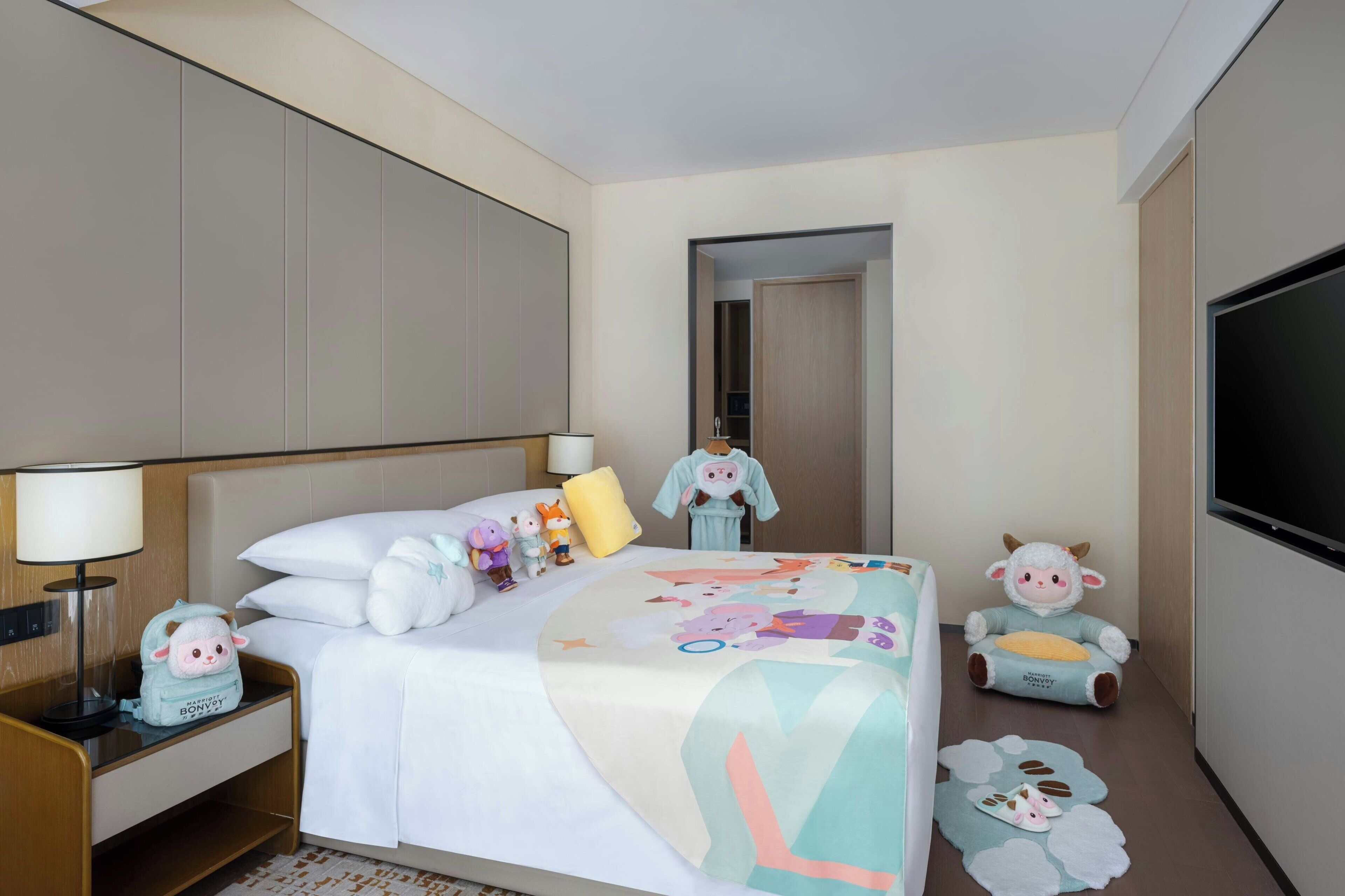 Una habitación de hotel con cama, mesita de noche, lámpara, peluches y televisor.
