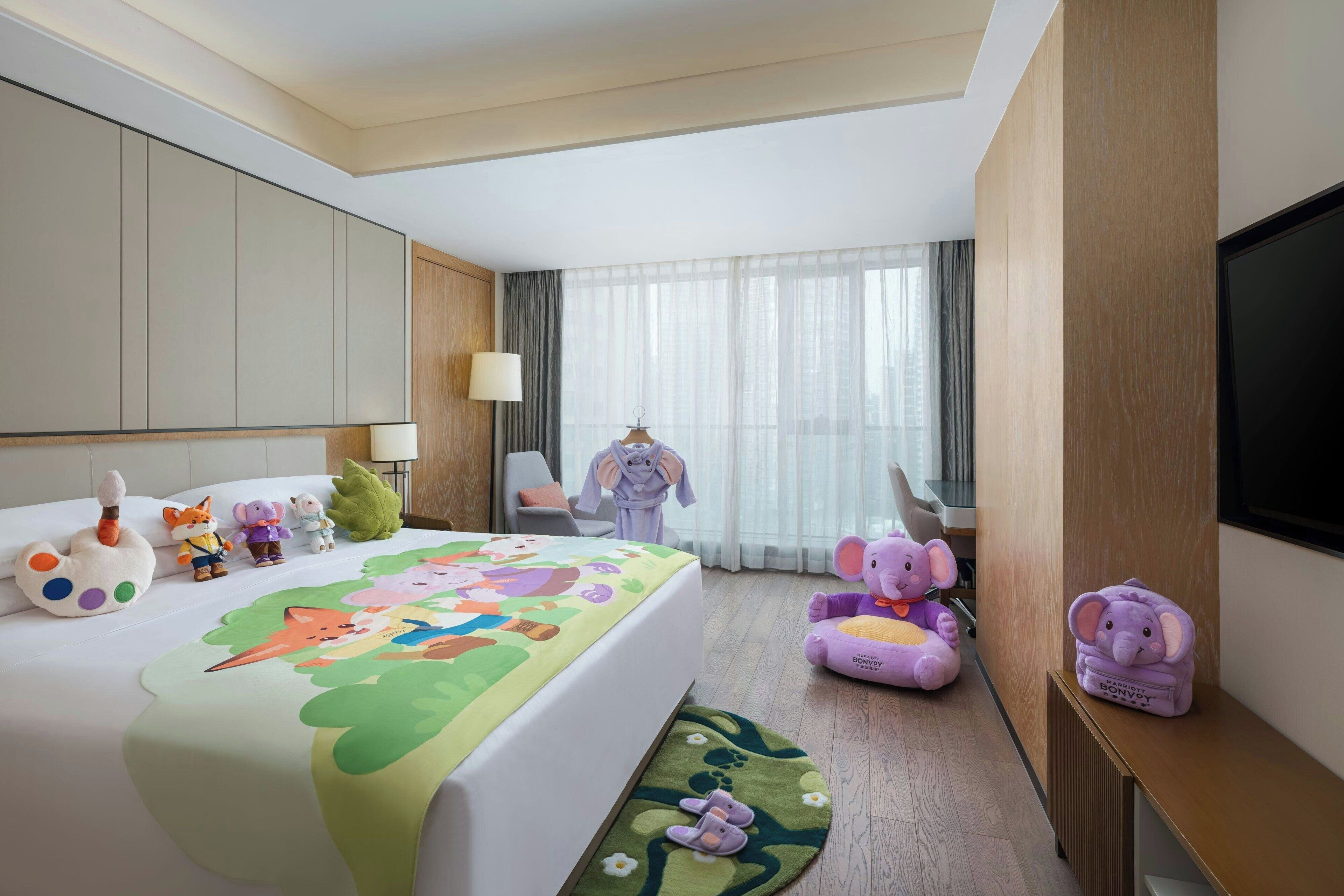 A hotel room with a bed, a desk, a chair, a TV, and various children's toys.